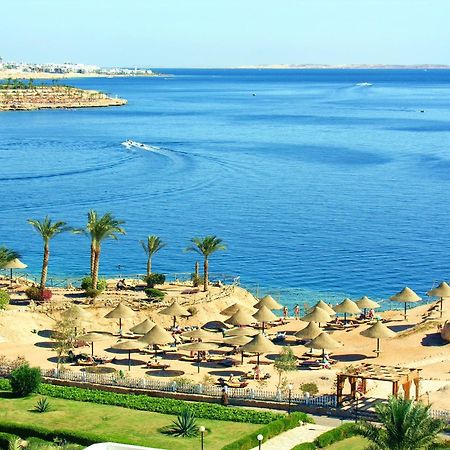 V Hotel Sharm El Shiekh Exterior photo The photo depicts a beautiful coastal scene featuring a serene blue sea and a sandy beach. There are several thatched umbrellas set up along the beach, suggesting a relaxing atmosphere for sunbathing or lounging. Palms are scattered throughout the ar
