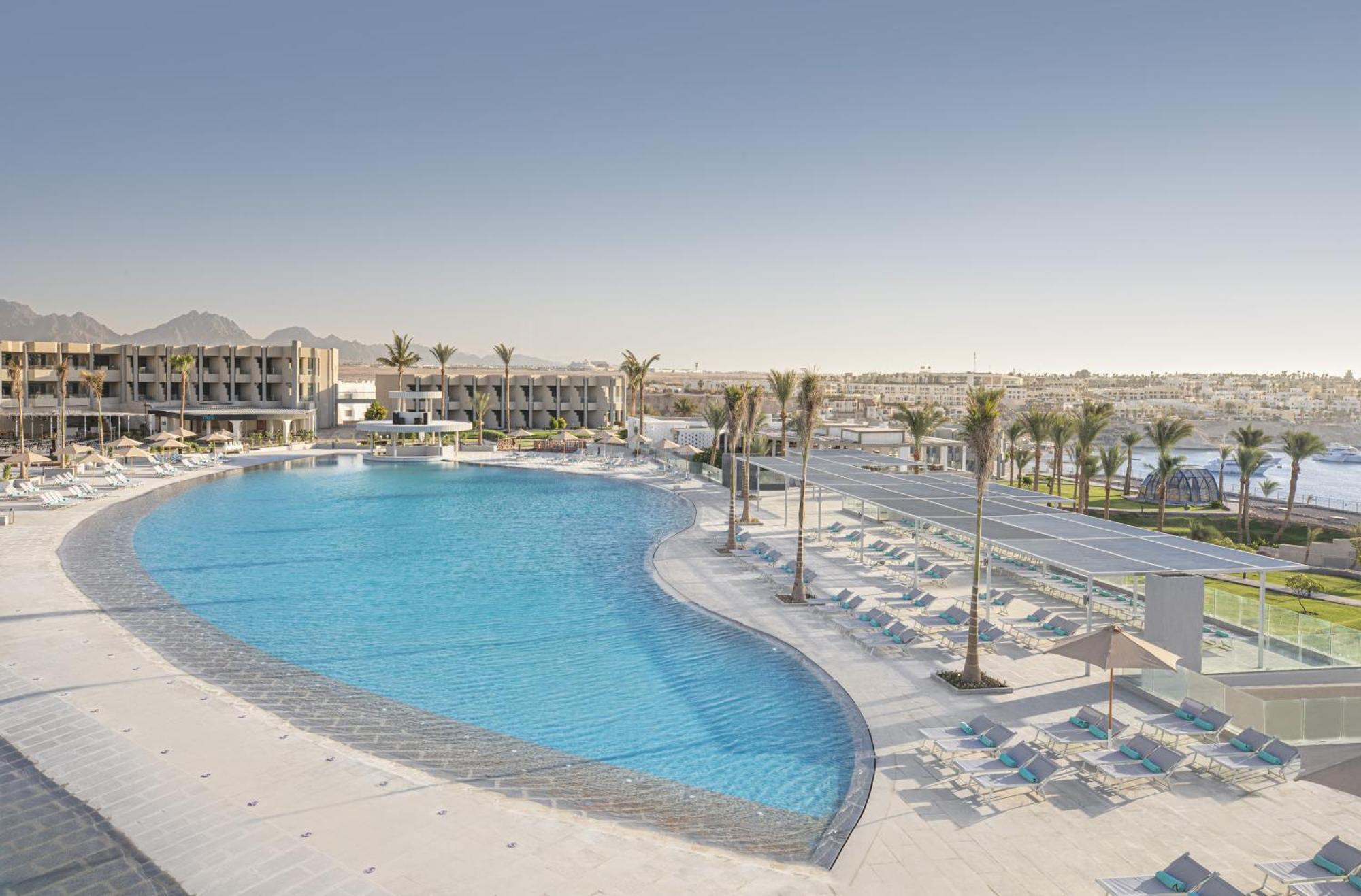 V Hotel Sharm El Shiekh Exterior photo The photo shows a large, inviting swimming pool with a curved shape, surrounded by sun loungers and palm trees. In the background, there are modern buildings that likely belong to a resort or hotel. The area appears to be sunny and well-maintained, w