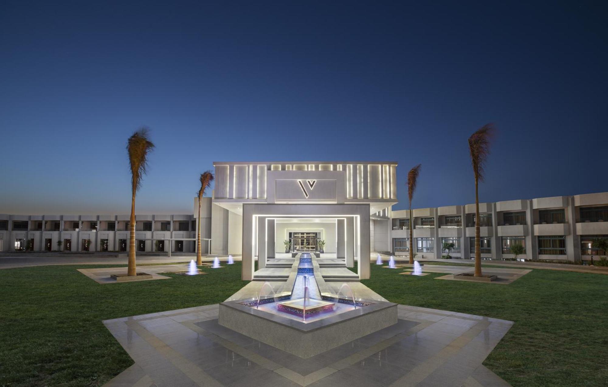 V Hotel Sharm El Shiekh Exterior photo The photo shows a modern architectural building illuminated at dusk. The structure features a central entrance framed by illuminated elements and has a sleek, contemporary design. In front of the building, there is a water feature with a fountain, wh