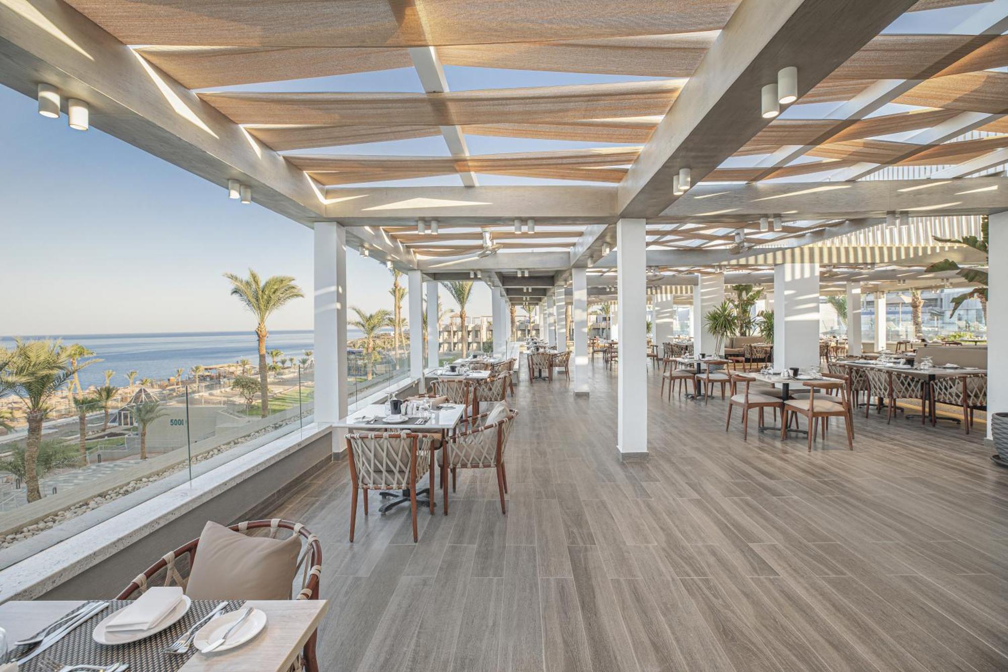 V Hotel Sharm El Shiekh Exterior photo The photo shows a stylish restaurant interior with a modern design. It features large windows that provide a panoramic view of the beach or ocean beyond. The space is bright and airy, enhanced by wooden beams on the ceiling and ample natural light. T
