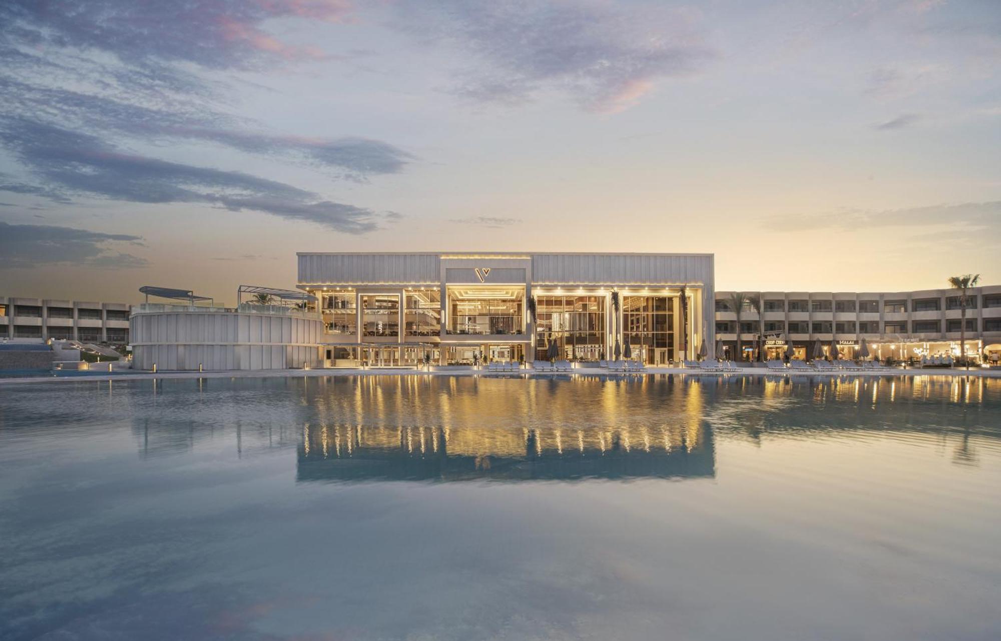 V Hotel Sharm El Shiekh Exterior photo The photo depicts a modern architectural building situated near a body of water, likely a lake or a pond. The structure features large glass windows, allowing for a bright and airy interior. The setting appears tranquil, with a beautiful sunset or tw