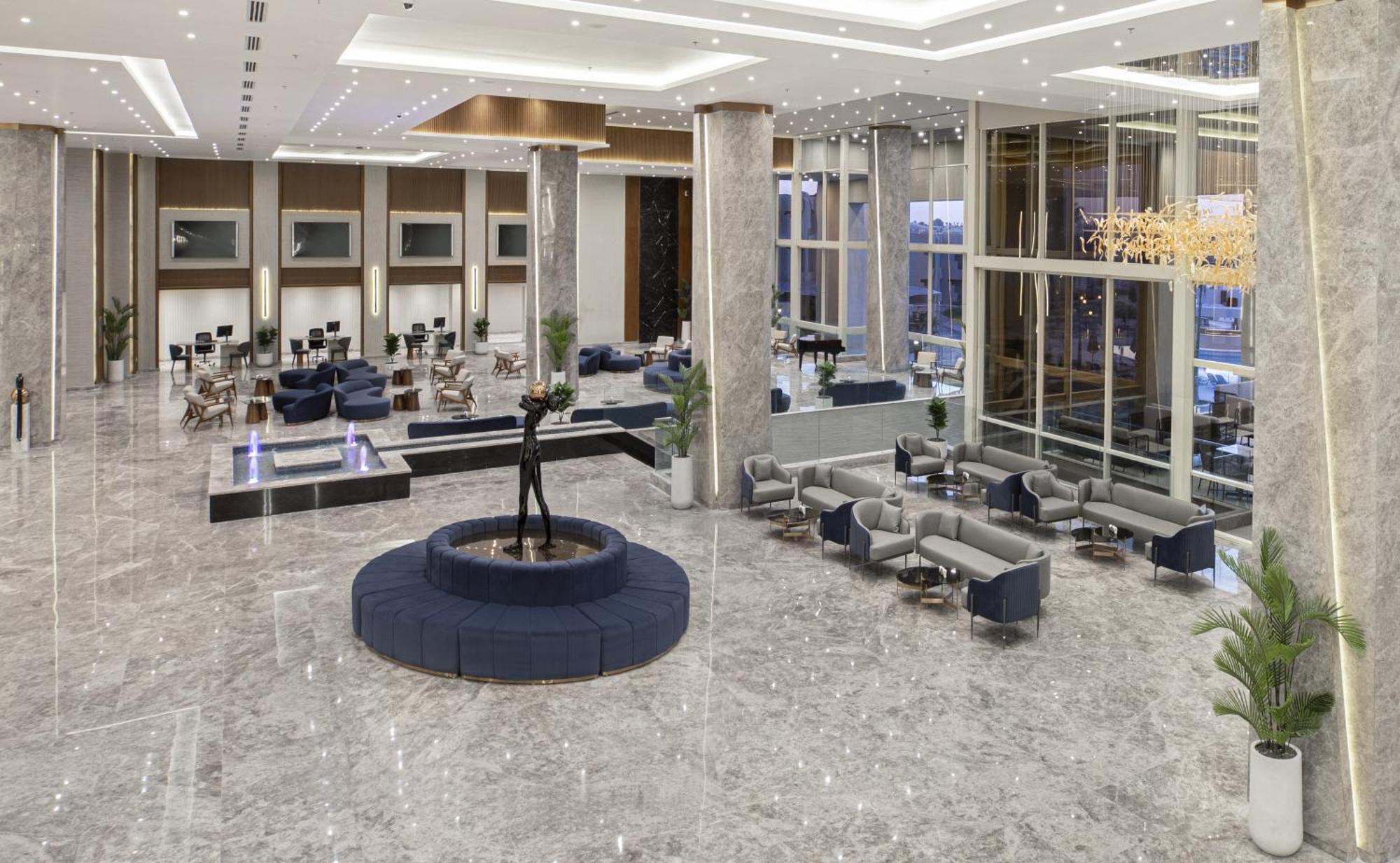 V Hotel Sharm El Shiekh Exterior photo The photo depicts a modern and spacious hotel lobby. The floor is made of polished marble, giving it an elegant look. In the center, there is a circular seating area with a fountain, surrounded by stylish blue and gray chairs. Large windows allow nat