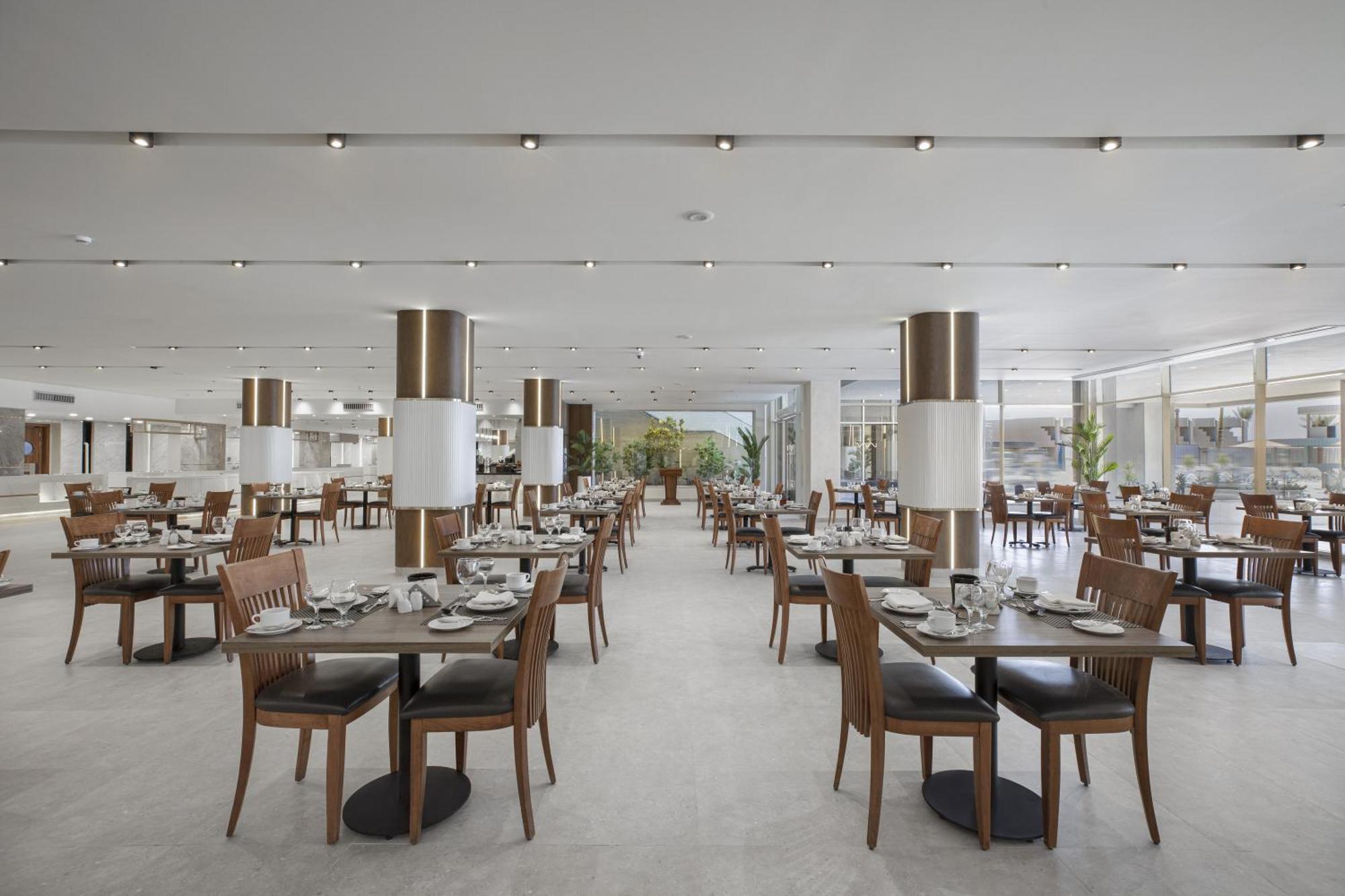 V Hotel Sharm El Shiekh Exterior photo The photo depicts a restaurant interior. The space features a modern design with several rows of wooden tables and chairs, neatly arranged for diners. Each table is set with dishes and cutlery, indicating readiness for service. Bright lighting is pro