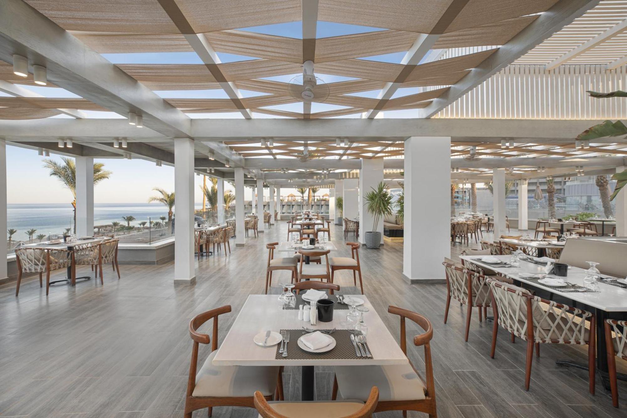 V Hotel Sharm El Shiekh Exterior photo The photo shows the interior of a modern restaurant with a spacious, open design. The ceiling features a geometric pattern of beams and fabric, allowing natural light to filter through. The dining area consists of several tables with wooden chairs, n