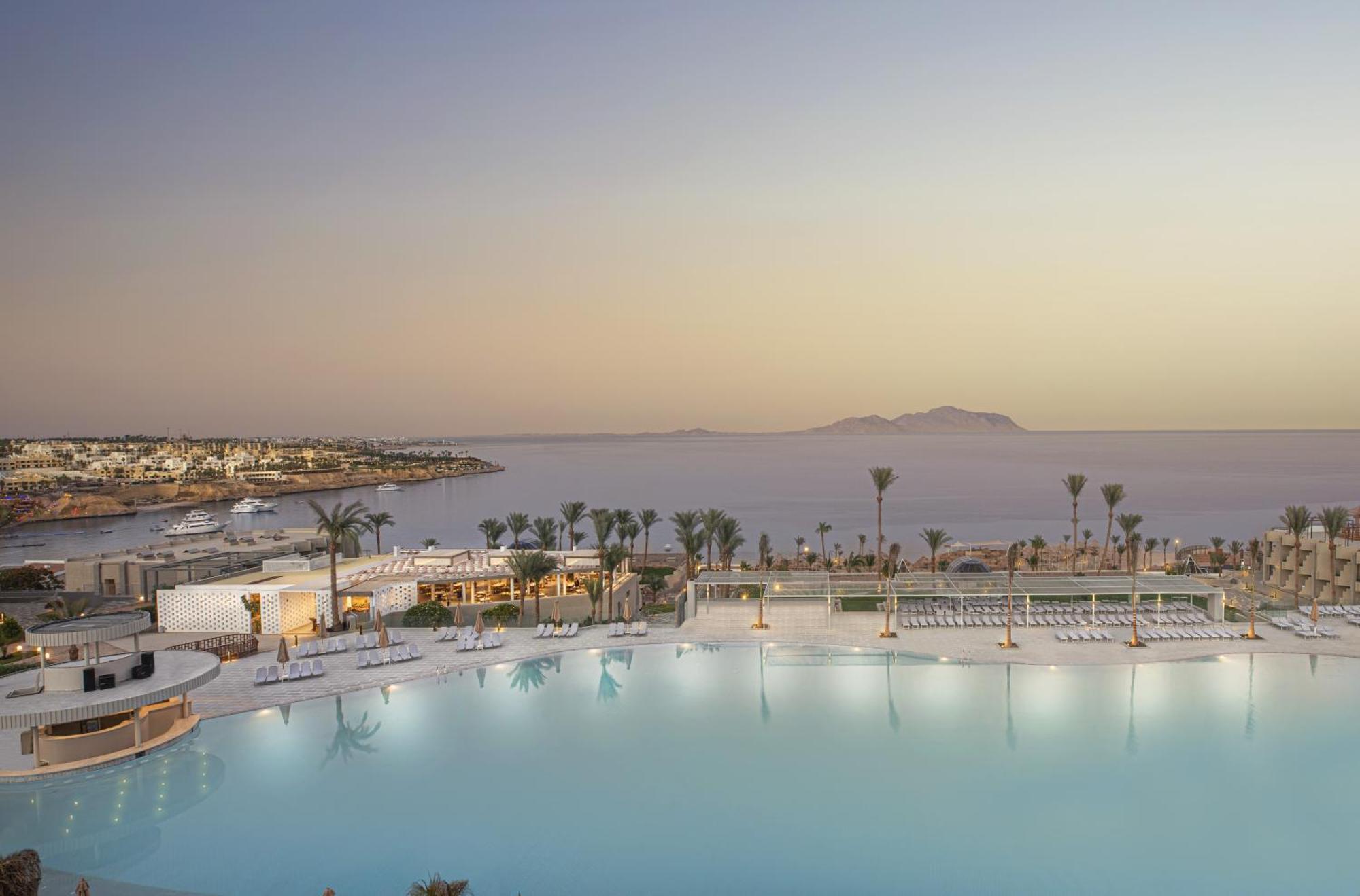 V Hotel Sharm El Shiekh Exterior photo The photo showcases a beautiful resort setting featuring a large swimming pool that reflects the sky. Surrounding the pool are lounge chairs and palm trees, adding a tropical feel. In the background, there’s a view of the coastline and distant mounta