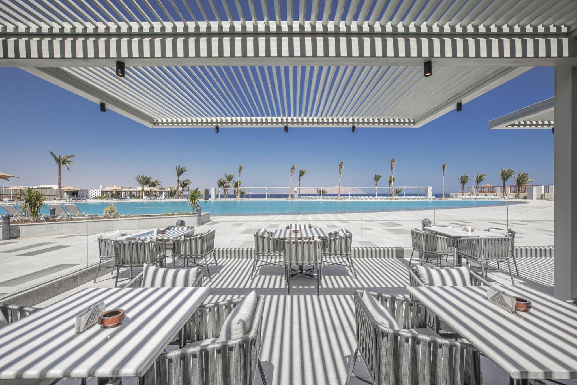 V Hotel Sharm El Shiekh Exterior photo The photo shows a stylish outdoor dining area, featuring tables and chairs with striped patterns. There are large, adjustable overhead awnings creating alternating shadows on the patio. In the background, a large, tranquil pool reflects the clear blu