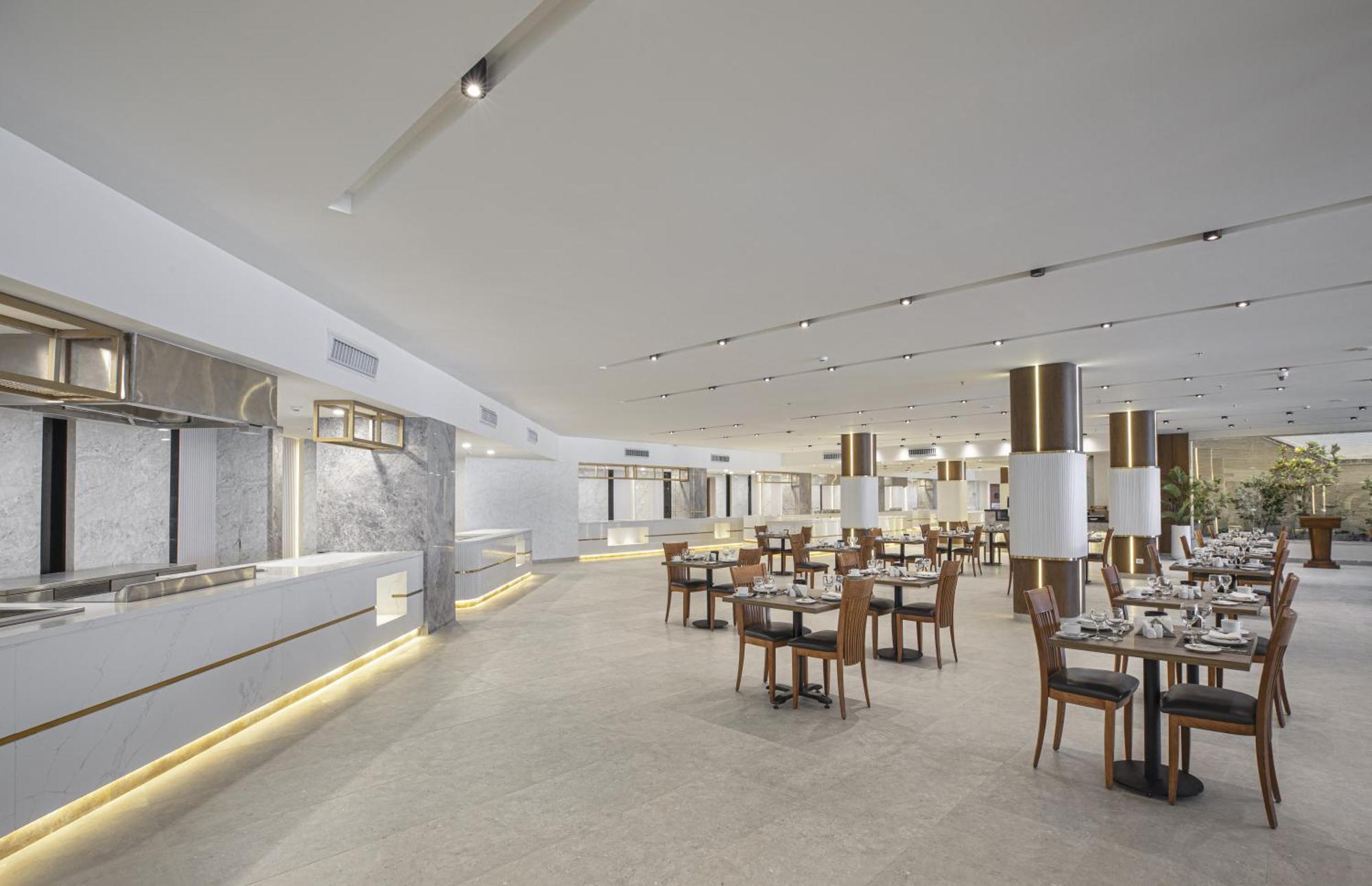 V Hotel Sharm El Shiekh Exterior photo The photo depicts a modern restaurant interior. The space features a bright and airy ambiance with large windows and an open layout. There are several tables arranged neatly across the floor, surrounded by wooden chairs. The decor includes light-colo