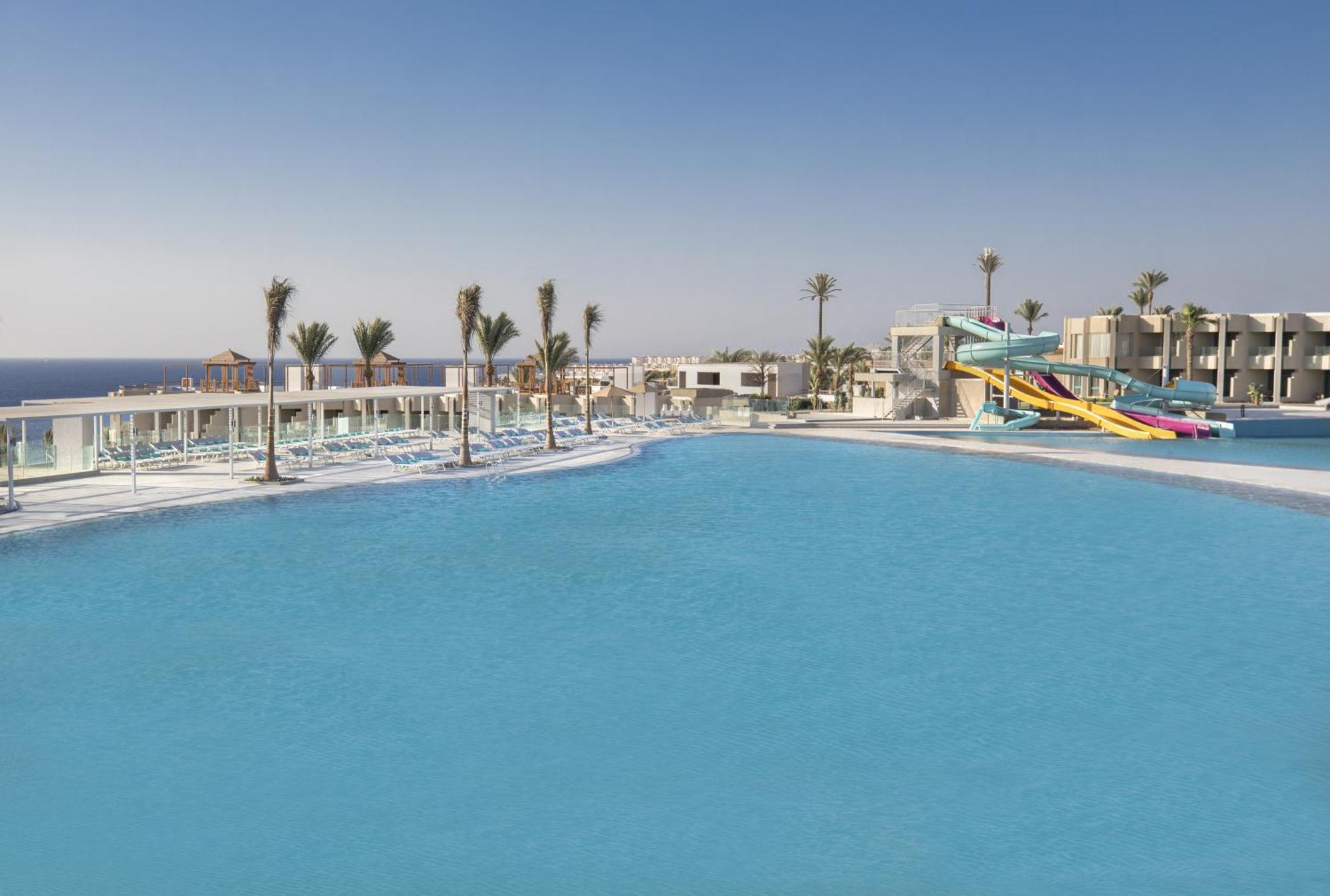 V Hotel Sharm El Shiekh Exterior photo The photo shows a sunny outdoor pool area with clear blue water. In the foreground, there is a large swimming pool, and in the background, several palm trees line the edges. To the right, a colorful water slide is visible, suggesting a fun recreation