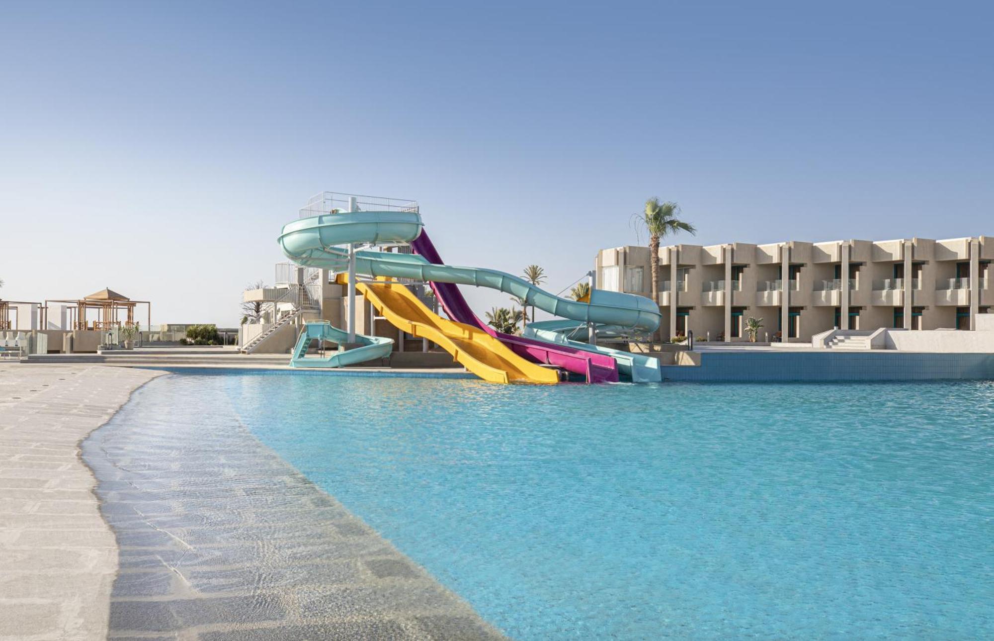 V Hotel Sharm El Shiekh Exterior photo The photo depicts a vibrant swimming pool area featuring two large water slides. One slide is turquoise and the other is yellow with purple accents. The pool is expansive, with clear blue water, and there are palm trees in the background, suggesting 