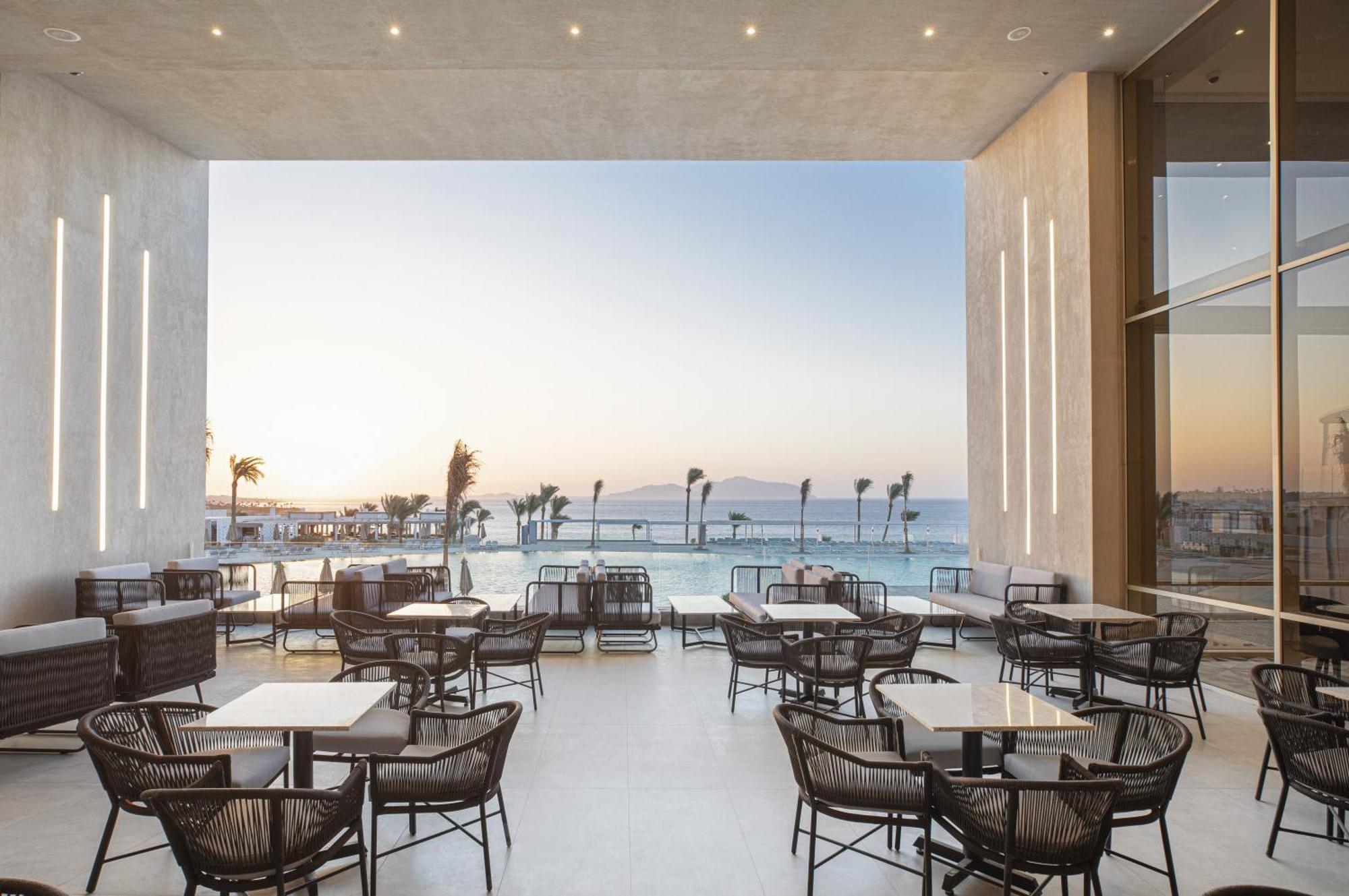 V Hotel Sharm El Shiekh Exterior photo The photo depicts a modern indoor dining area with several wicker chairs and tables arranged neatly. Large glass panels create an open space that offers a scenic view of the sea and a sunset in the background, where gentle colors of orange and blue b