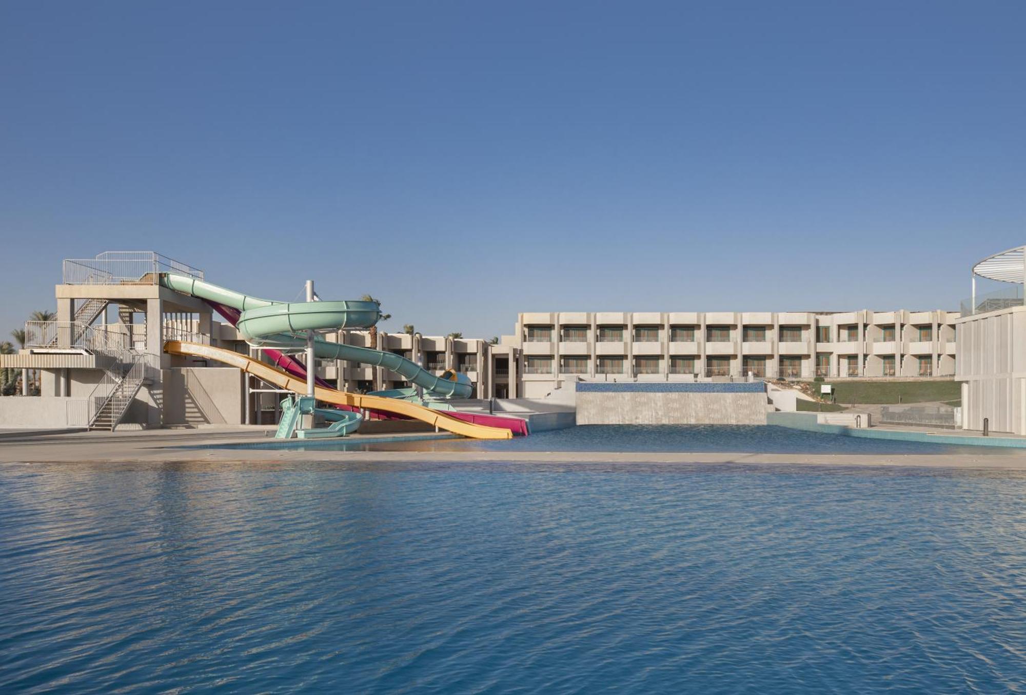 V Hotel Sharm El Shiekh Exterior photo The photo shows a swimming pool area featuring a large water slide with multiple colorful lanes. The slide is attached to a raised structure. In the background, there is a building with multiple stories, likely serving as accommodations or facilities