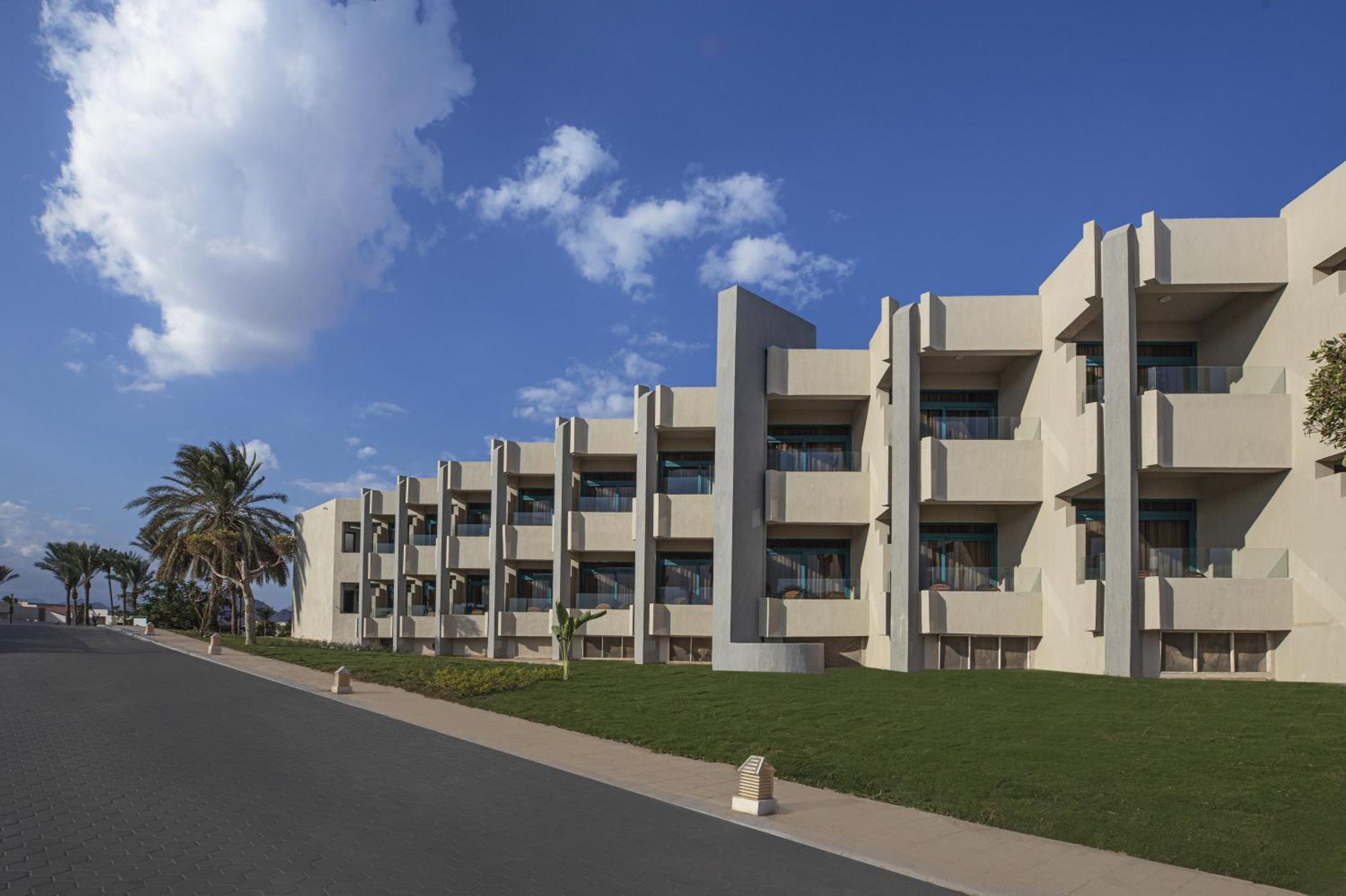 V Hotel Sharm El Shiekh Exterior photo The photo shows a modern building with multiple stories, featuring a streamlined architectural design. It has several balconies or terraces, and large windows that suggest a bright and open environment. The building is set against a backdrop of a blu
