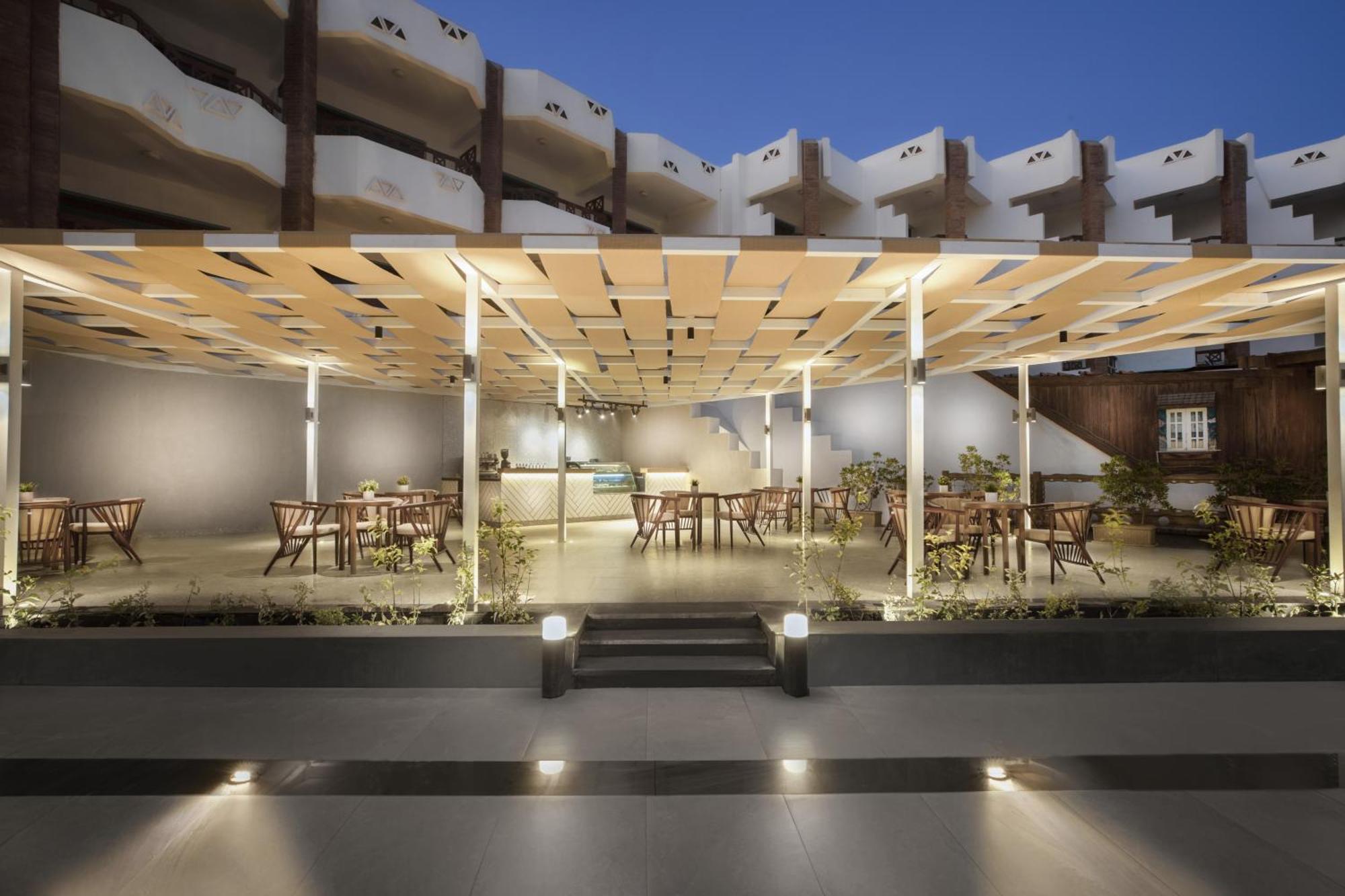 V Hotel Sharm El Shiekh Exterior photo The photo depicts a modern outdoor dining area or café setting. The space features a structured canopy made of grid-like panels, providing shade. Below the canopy, there are several wooden tables and chairs arranged neatly. The area is well-lit, with