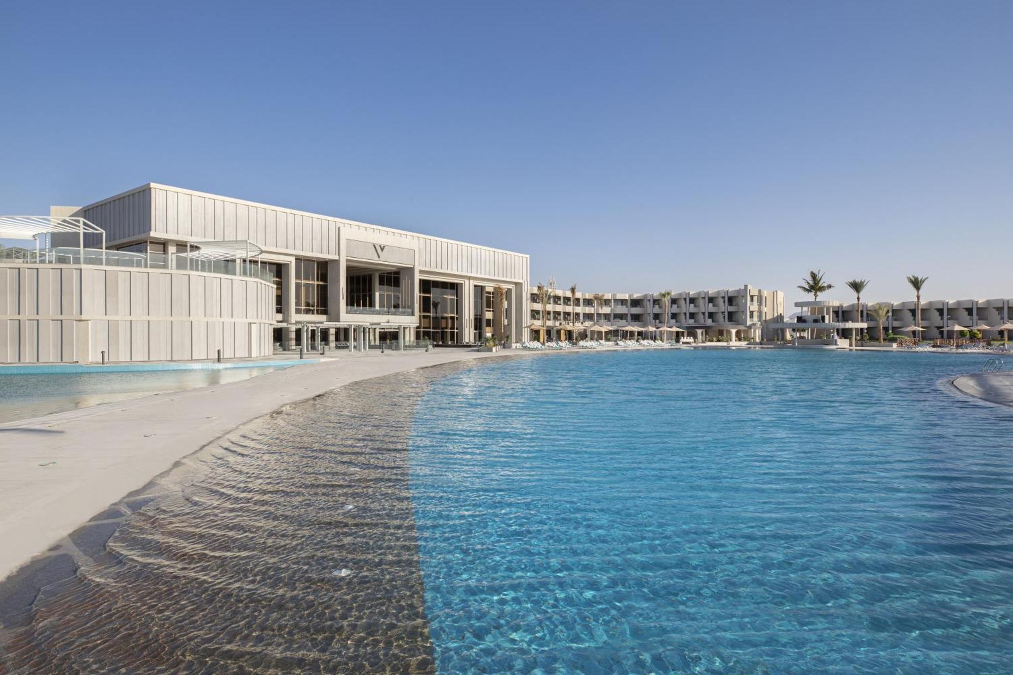 V Hotel Sharm El Shiekh Exterior photo The photo shows a modern hotel or resort building situated near a large swimming pool. The pool has a gentle, wavy edge and is filled with clear blue water. In the background, there are multiple buildings that appear to be part of the same resort com