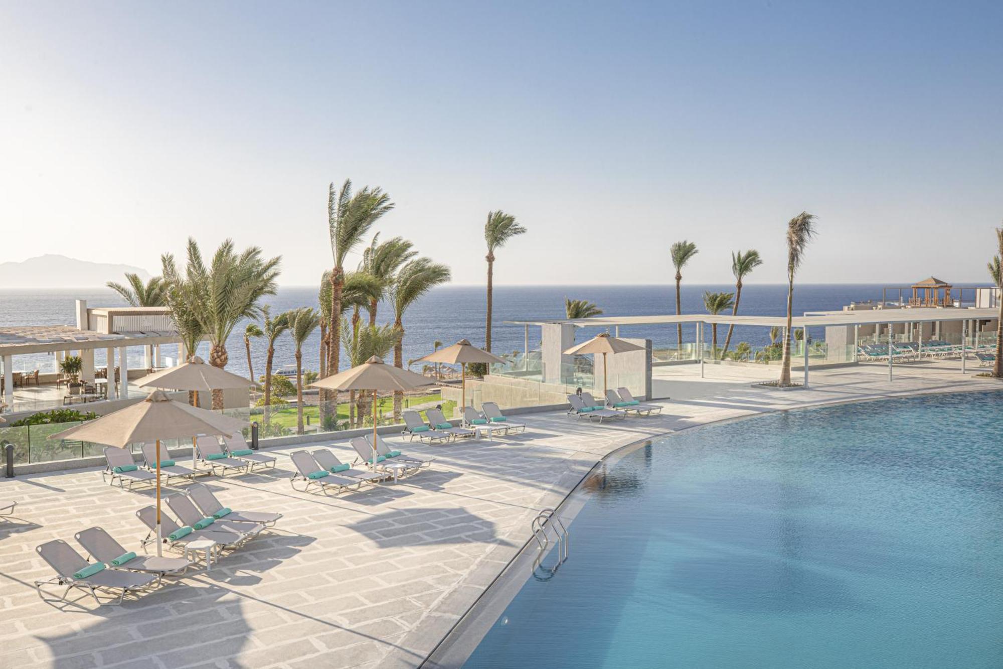 V Hotel Sharm El Shiekh Exterior photo The photo shows a tranquil poolside scene at a resort. There are several sun loungers arranged around a large swimming pool. Palm trees are visible, swaying gently in the breeze, and the background features a beautiful view of the ocean. The atmosphe