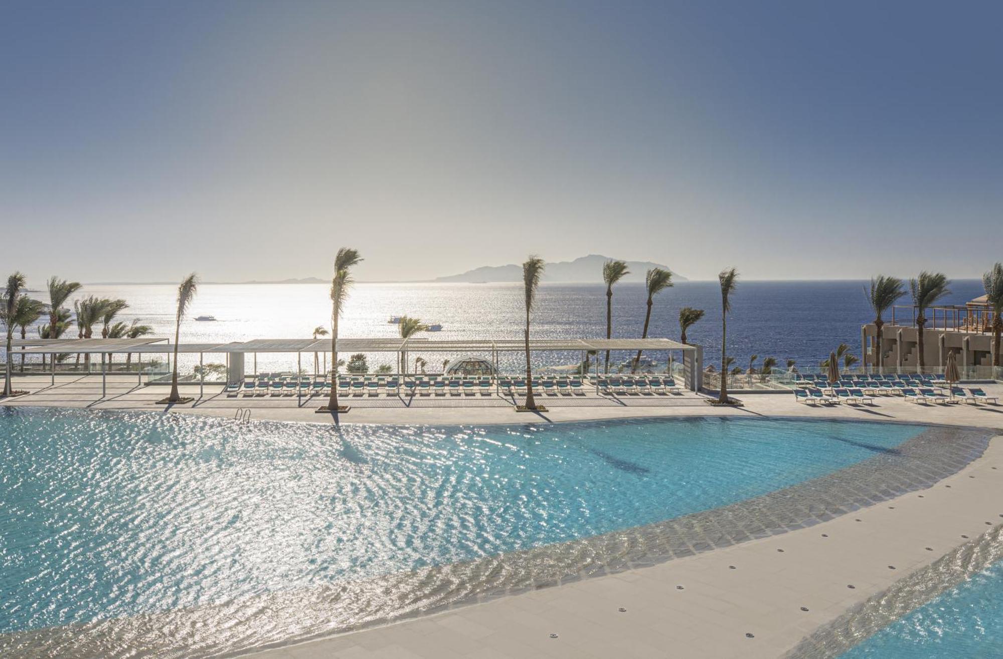 V Hotel Sharm El Shiekh Exterior photo The photo shows a serene pool area overlooking the ocean. The pool is large and features a sleek design with a gentle curve. Surrounding the pool are lounge chairs arranged for relaxation. In the background, there are palm trees swaying gently, sugge
