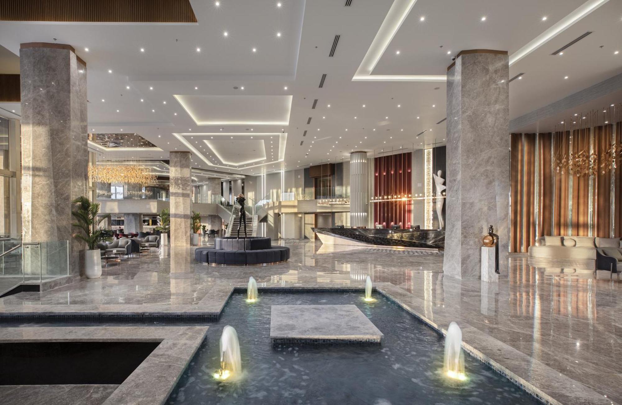 V Hotel Sharm El Shiekh Exterior photo The photo shows the interior of a modern hotel lobby. The space features an open layout with polished marble flooring and large pillars. Soft lighting illuminates the area, creating a warm ambiance. In the center, there is a water feature with small 