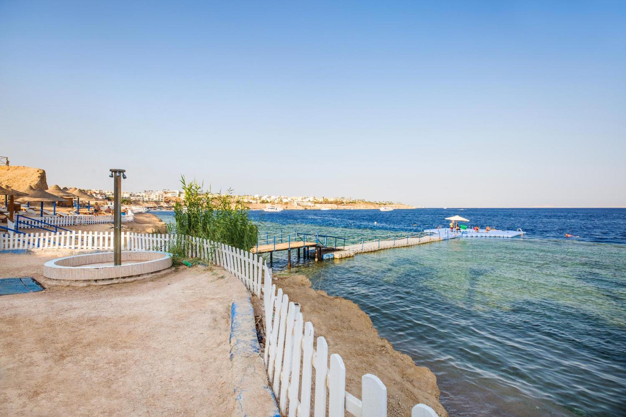 V Hotel Sharm El Shiekh Exterior photo The photo depicts a serene beach scene with clear blue water and a sandy shoreline. In the foreground, there is a circular structure that appears to be a small seating area or hot tub, surrounded by a white picket fence. The beach extends along the w