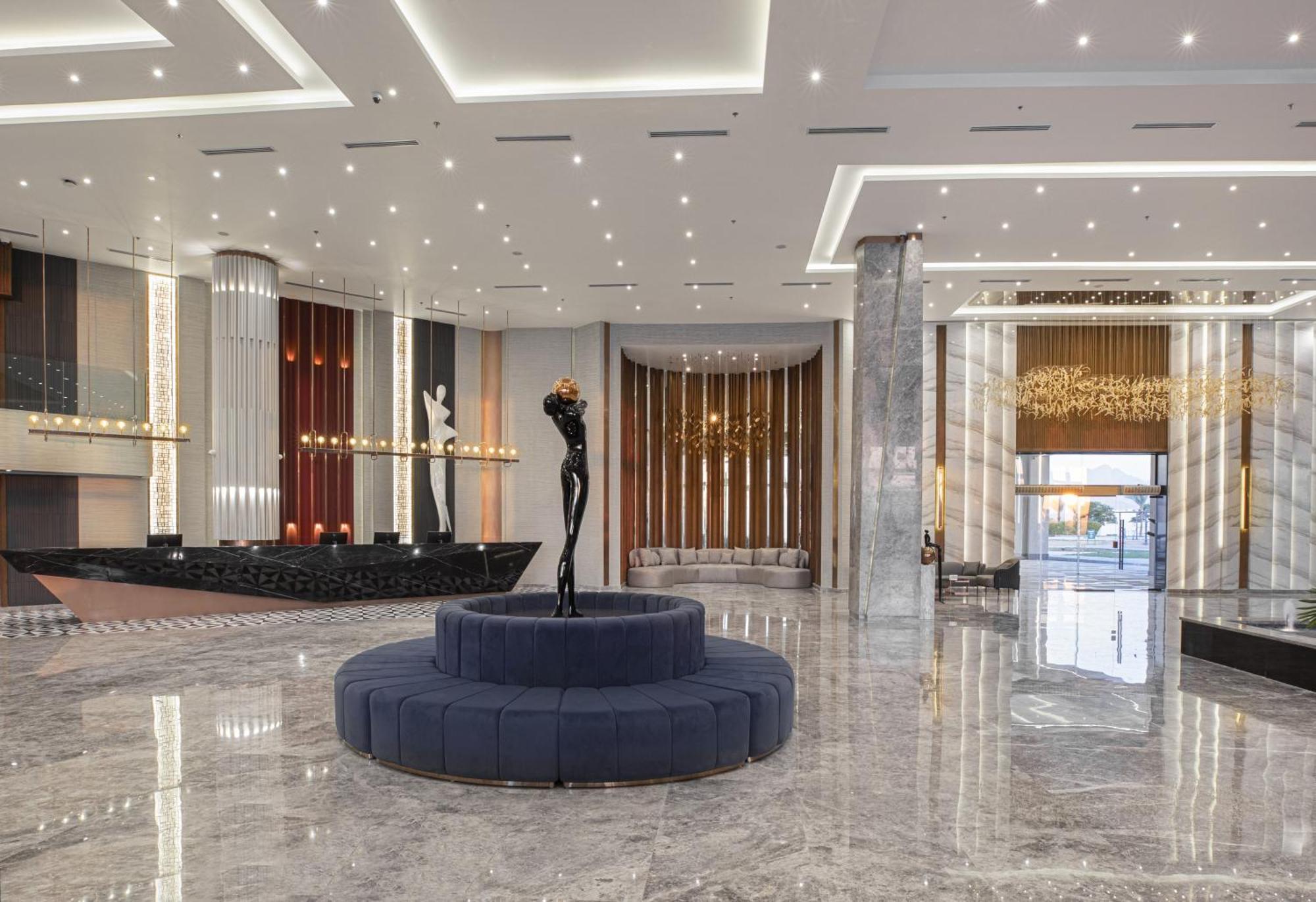 V Hotel Sharm El Shiekh Exterior photo The photo displays a modern hotel lobby with a sleek and elegant design. The flooring is made of polished marble, reflecting the ambient light. In the center, there is a circular blue velvet seating area surrounded by a minimalist black sculpture. Th
