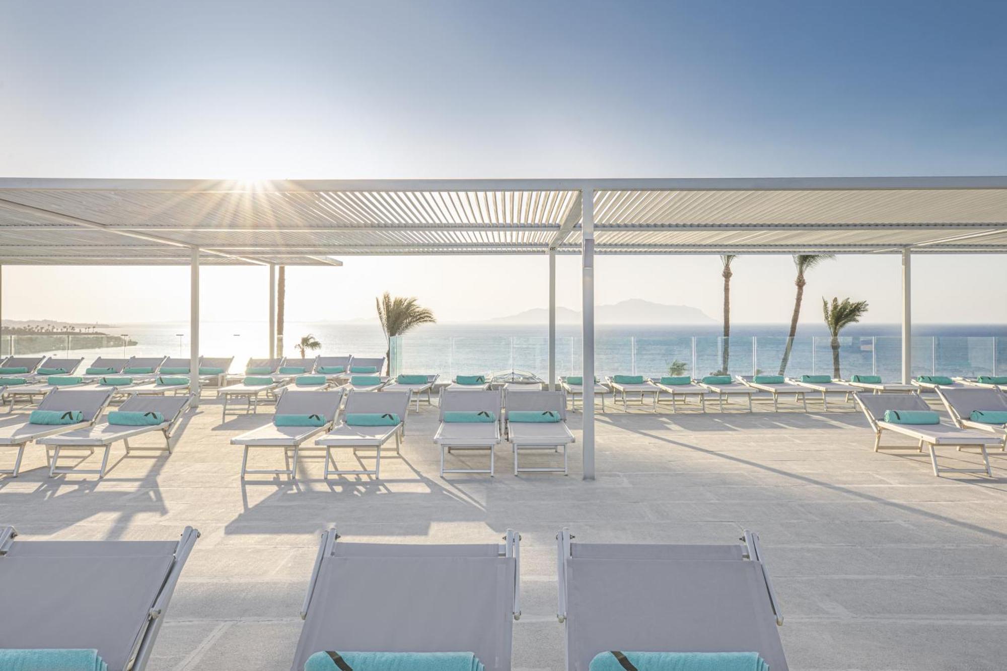 V Hotel Sharm El Shiekh Exterior photo The photo shows a serene outdoor space by the ocean, featuring numerous lounge chairs arranged neatly under a shaded structure. The chairs have light-colored cushions, and the area is bathed in soft sunlight, suggesting a warm and inviting atmosphere