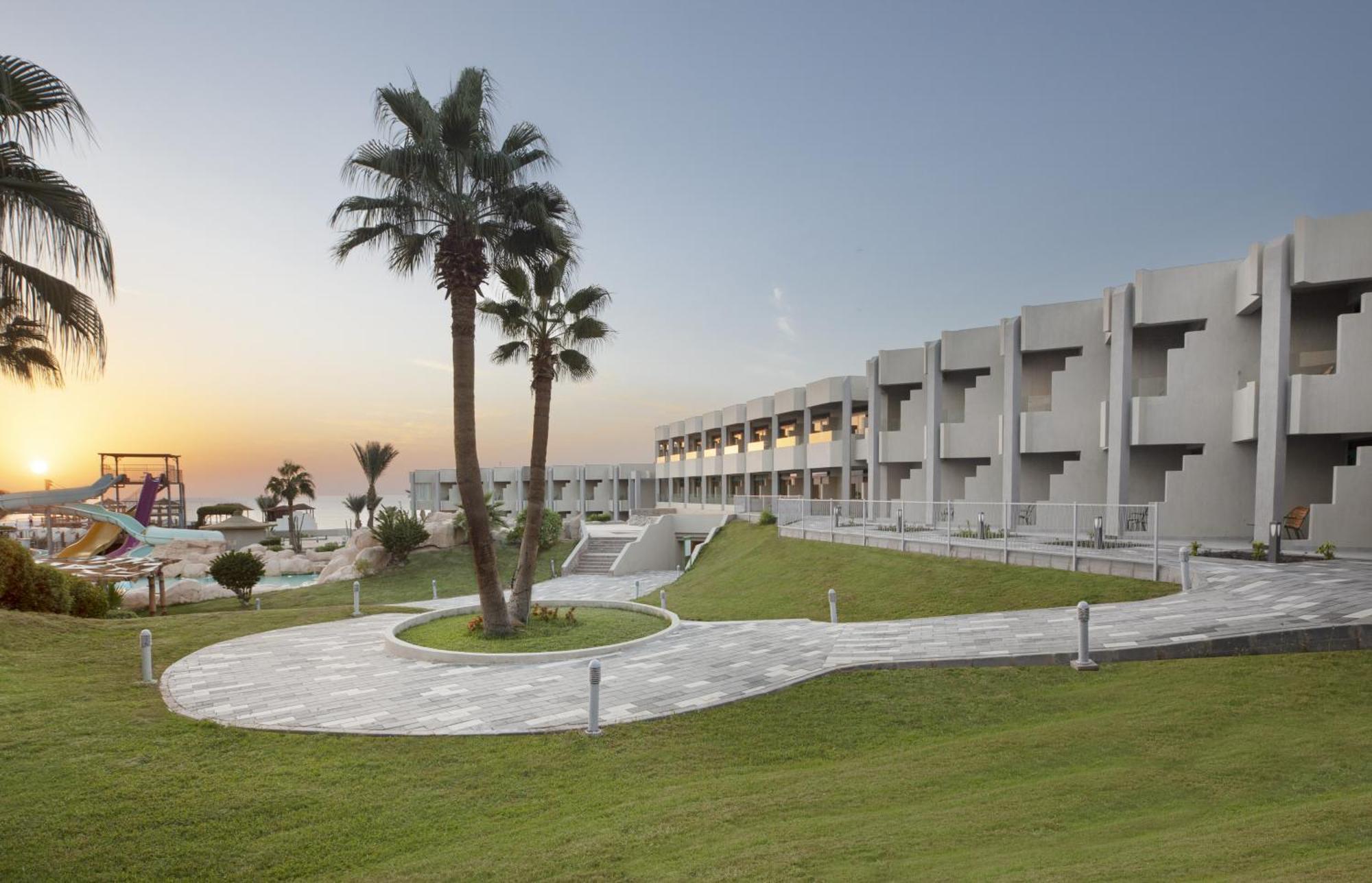 V Hotel Sharm El Shiekh Exterior photo The image depicts a modern resort or hotel complex set against a sunset backdrop. The architecture features a series of balconies and rooms that are likely part of the accommodation. In front of the building, there's a neatly landscaped lawn with a c