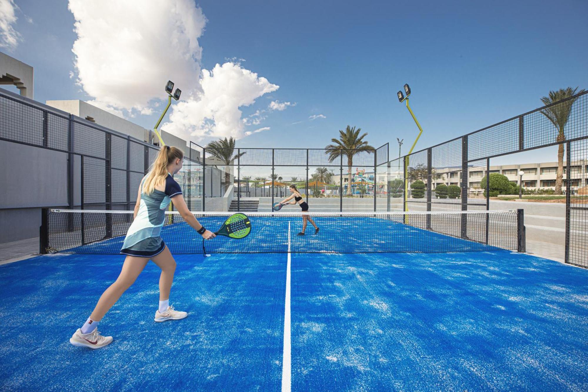 V Hotel Sharm El Shiekh Exterior photo The photo shows a sports scene on a blue padel tennis court. There are two players: one is in the foreground, preparing to hit a ball with a racket, and the other is in the background, getting ready to receive the shot. The court is enclosed with net