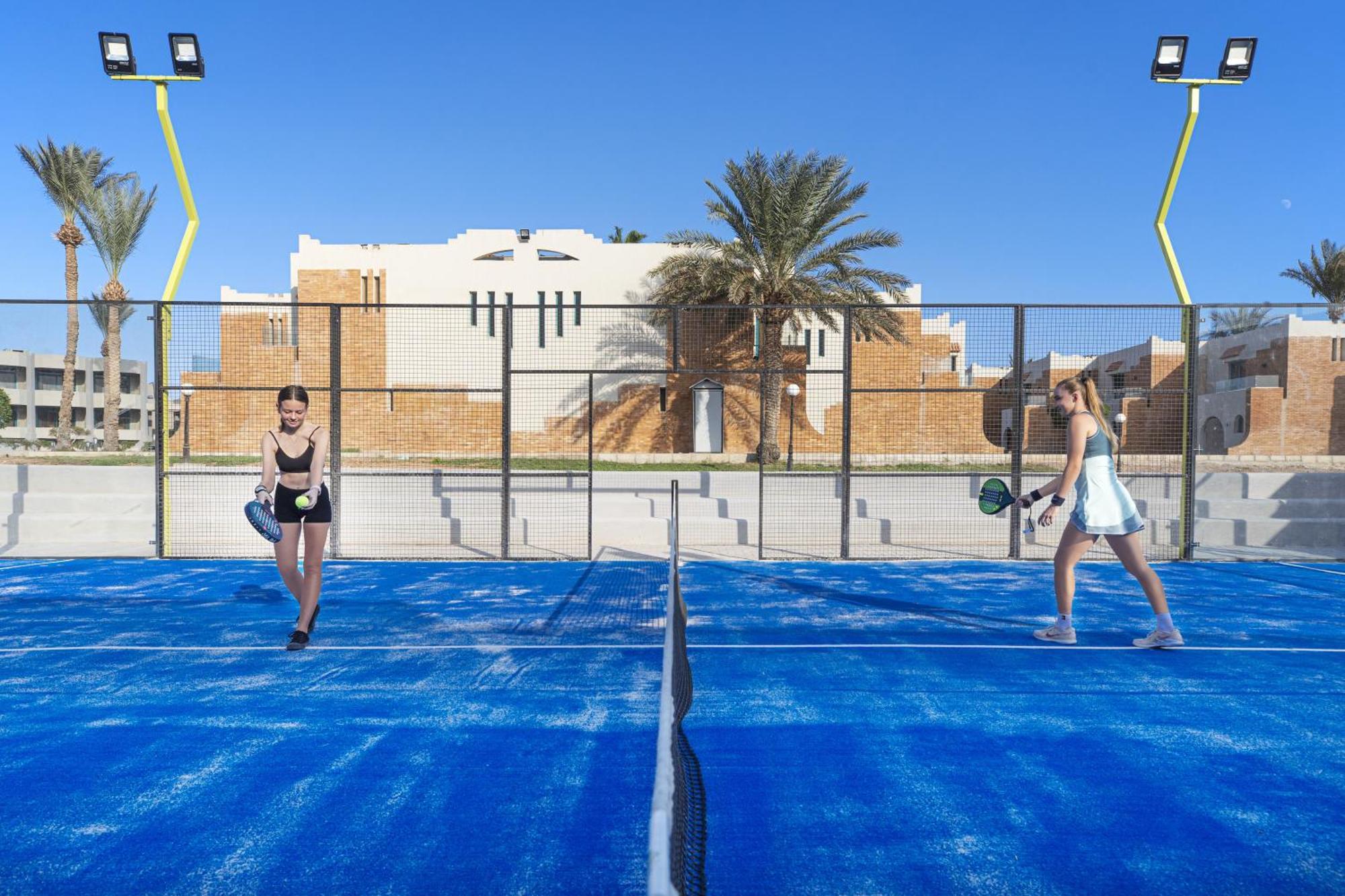 V Hotel Sharm El Shiekh Exterior photo The photo shows two women playing a racket sport on a blue court. They appear to be engaged in a match, possibly paddle tennis or a similar game. The background features a sunny sky and a building with a modern design, framed by palm trees. The court