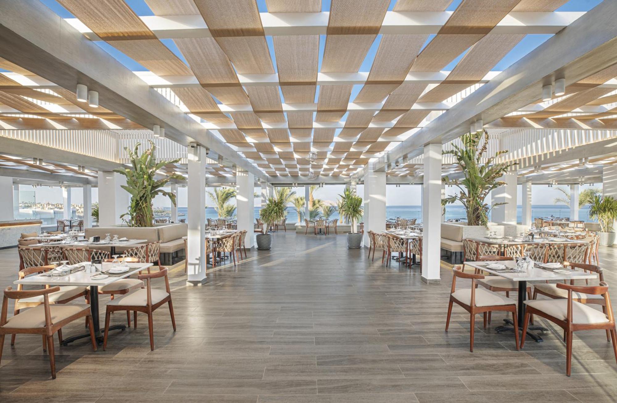 V Hotel Sharm El Shiekh Exterior photo The photo shows a spacious and modern restaurant interior with a bright and airy ambiance. The ceiling features a grid-like design with wooden slats, allowing natural light to filter through. There are several tables arranged neatly, surrounded by co