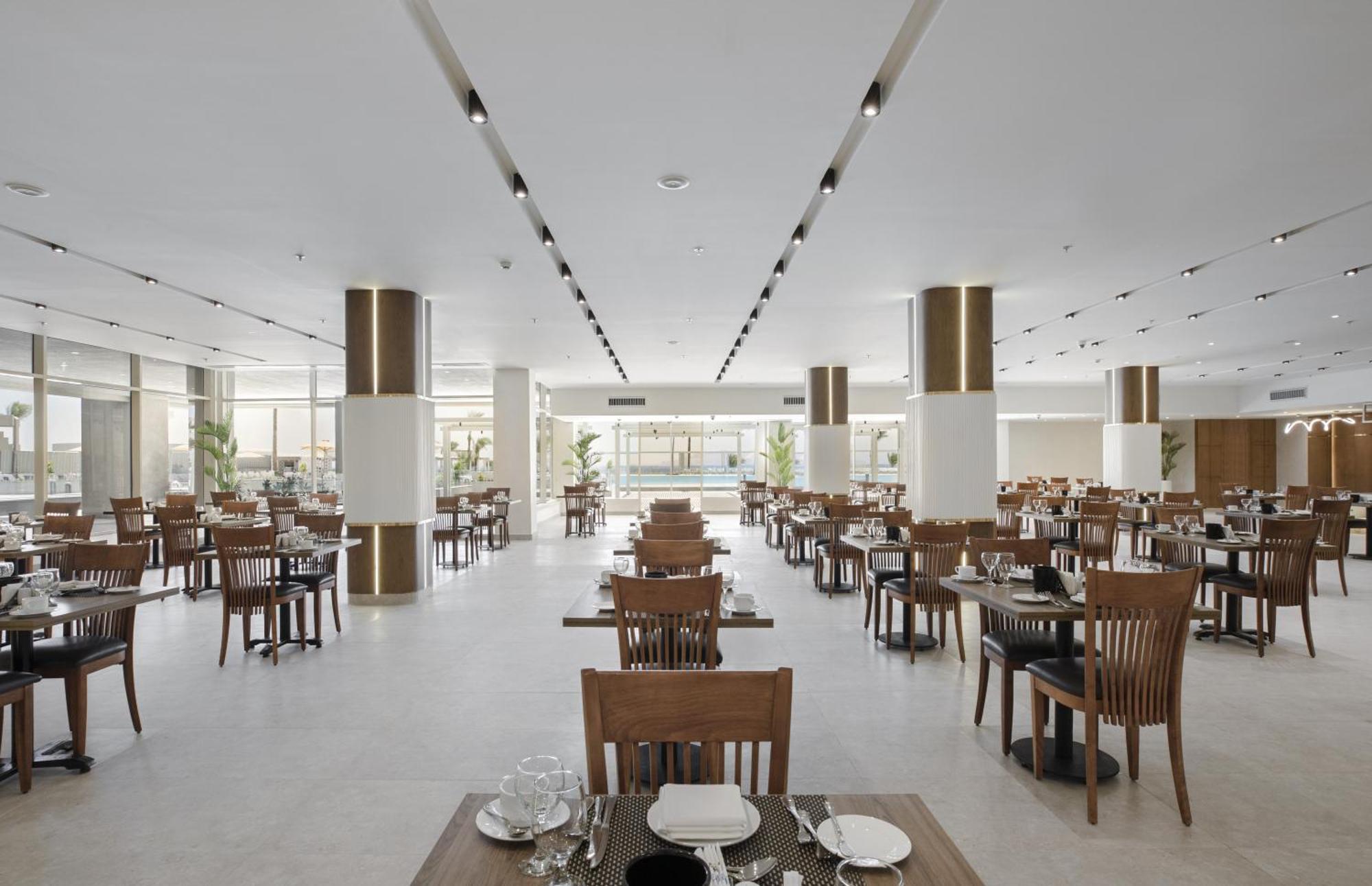 V Hotel Sharm El Shiekh Exterior photo The photo shows a spacious and modern restaurant interior. It features neatly arranged wooden tables with chairs and white table settings, including plates and cutlery. The design is minimalist with light-colored walls and polished flooring. There ar