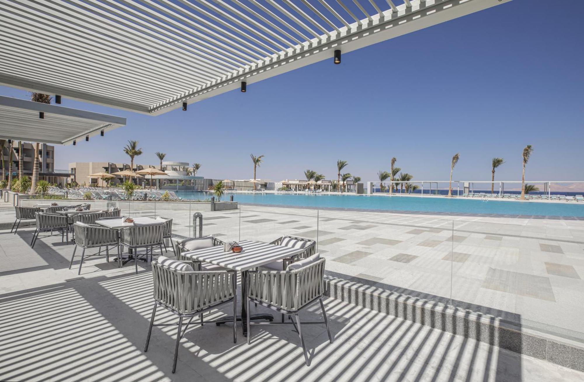 V Hotel Sharm El Shiekh Exterior photo The photo depicts a modern outdoor setting, likely at a resort or hotel. In the foreground, there are several stylish dining tables and chairs, arranged neatly under a shaded area created by slatted overhead structures. The background features a larg