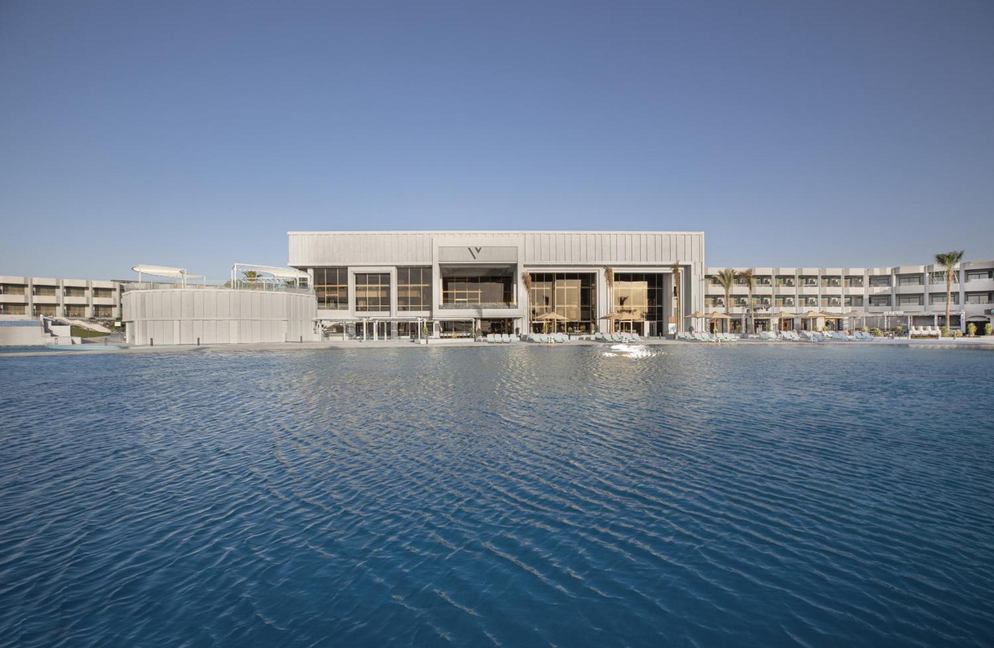 V Hotel Sharm El Shiekh Exterior photo The photo shows a modern building situated beside a large body of water. The architecture appears sleek and contemporary, with a facade that features a combination of glass and metallic elements. The scene is set against a clear blue sky, enhancing t