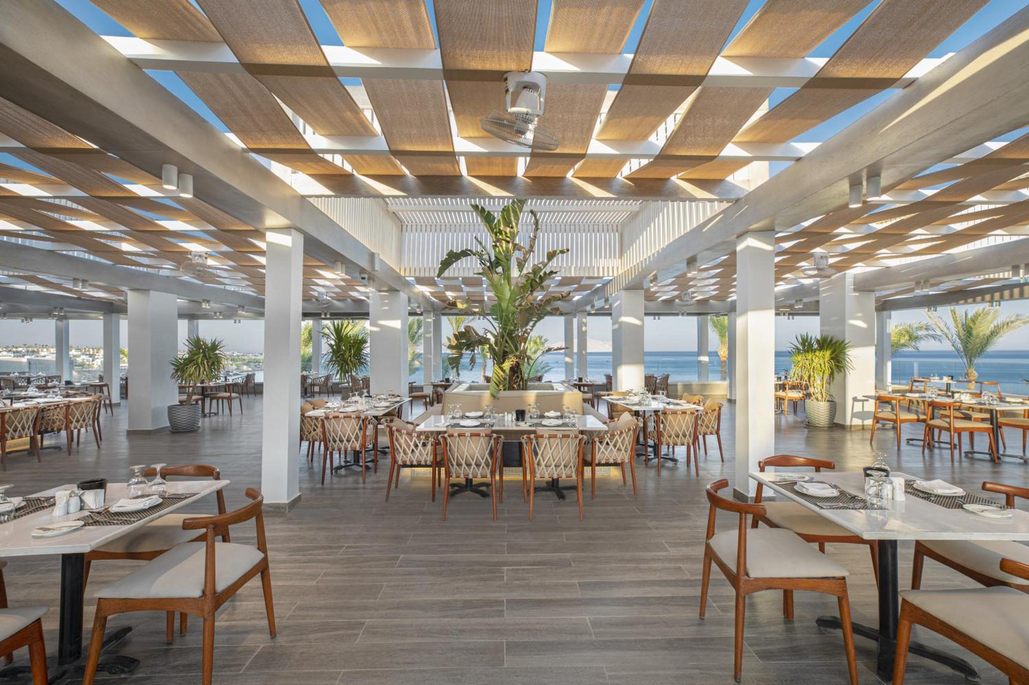 V Hotel Sharm El Shiekh Exterior photo The photo shows a spacious and modern restaurant interior. It features a bright and airy atmosphere with large windows letting in natural light. The ceiling has a series of wooden slats, creating a stylish pattern and providing shade. There's a centr