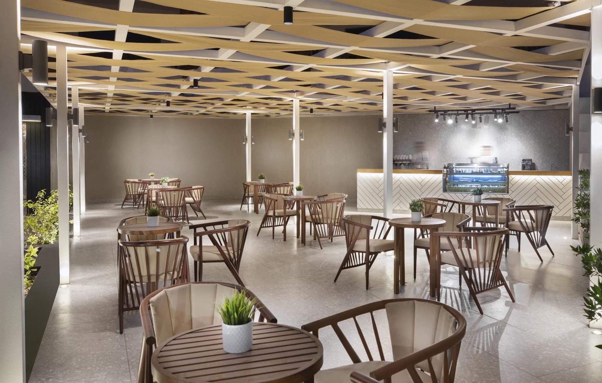 V Hotel Sharm El Shiekh Exterior photo The photo shows a modern indoor cafe or restaurant space. It features a series of wooden tables and chairs arranged throughout the area, with several plants placed on the tables. The ceiling has a distinctive geometric design with wooden beams, creat