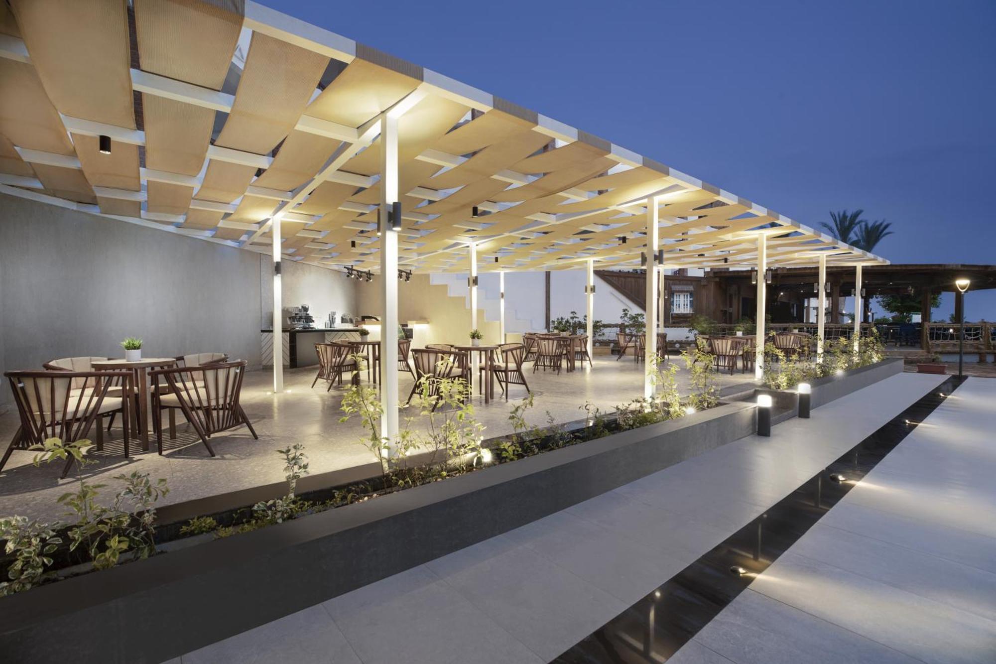 V Hotel Sharm El Shiekh Exterior photo The photo shows a modern outdoor dining area, featuring several wooden dining tables and chairs arranged under a stylish canopy. The canopy consists of slatted panels that allow light to filter through while providing some shade. The space is illumin