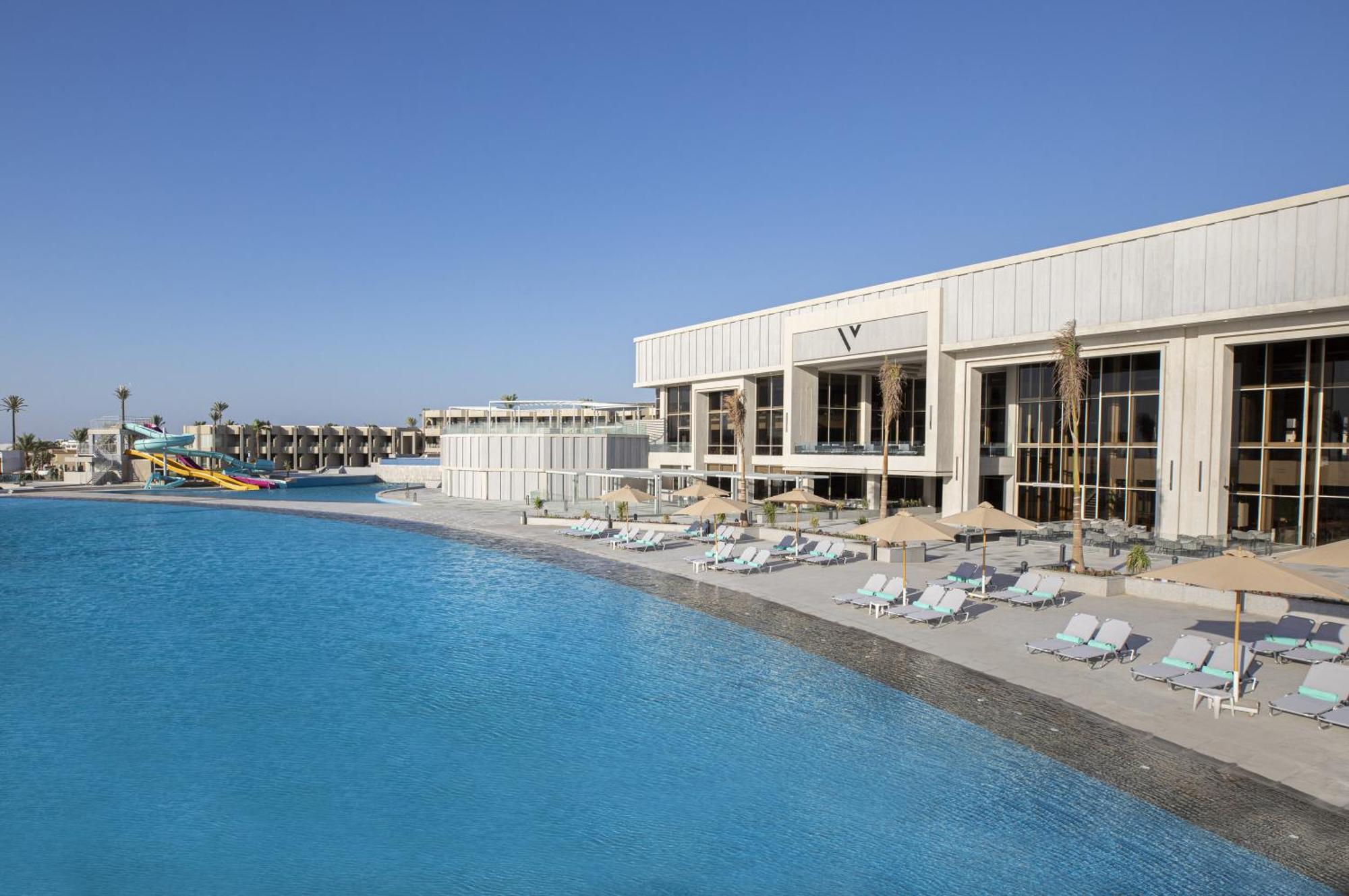 V Hotel Sharm El Shiekh Exterior photo The photo shows a large, tranquil swimming pool with crystal-clear water. Alongside the pool, there are several lounge chairs arranged for relaxation. In the background, there is a modern building with large glass windows, likely serving as a facilit