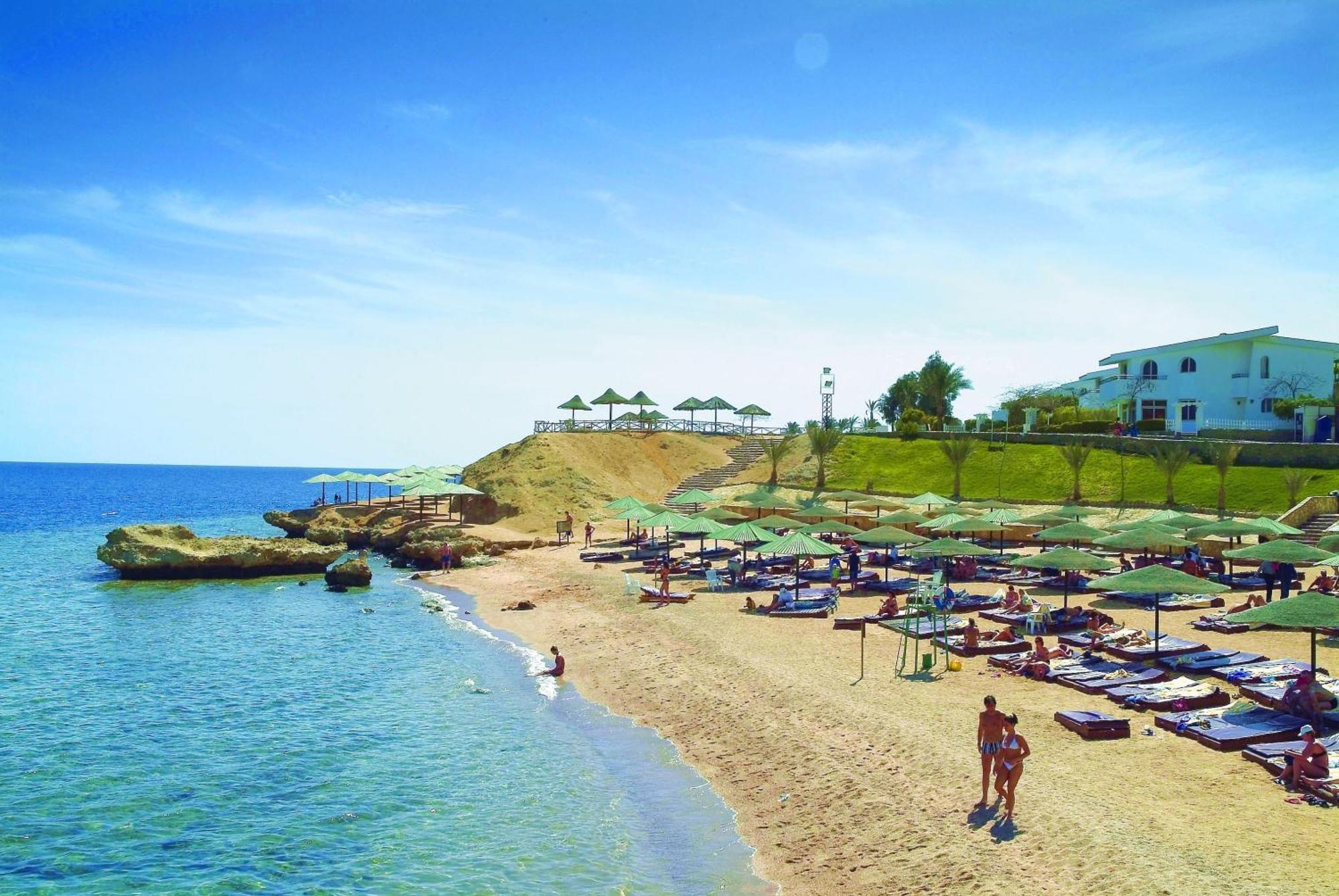 V Hotel Sharm El Shiekh Exterior photo The photo depicts a picturesque beach scene. There is a sandy shoreline with clear blue water gently lapping at the shore. Green umbrellas and lounge chairs are neatly arranged on the sand, providing shade for relaxation. In the background, there is 