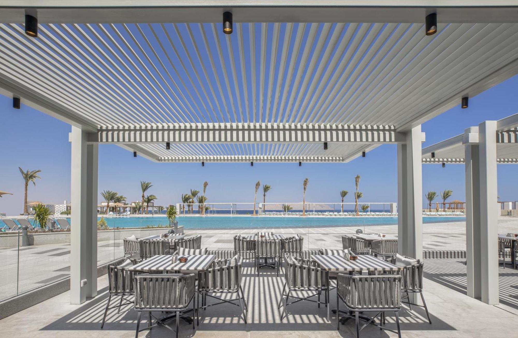 V Hotel Sharm El Shiekh Exterior photo The photo shows a modern outdoor dining area with a stylish pergola overhead. Beneath the pergola are several dining tables and chairs arranged in a neat layout. The setting appears to be near a swimming pool, with a clear blue sky and palm trees vis