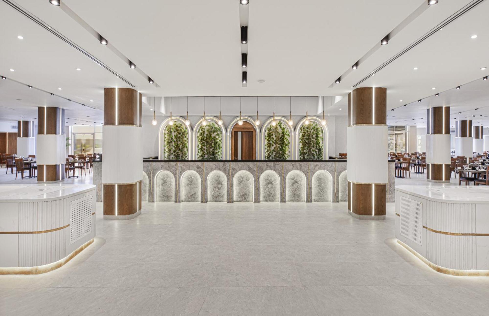 V Hotel Sharm El Shiekh Exterior photo The image depicts a modern restaurant interior. The space features a large, open design with a polished, light-colored floor. Prominent columns are styled with a combination of white and wood finishes. 

In the background, there is a stylish receptio