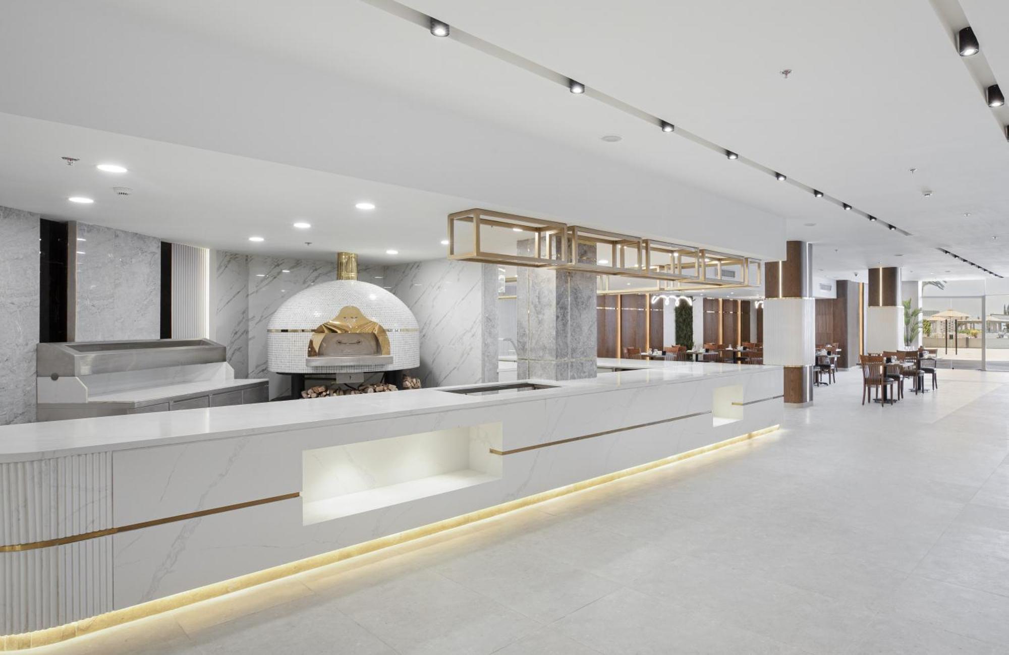V Hotel Sharm El Shiekh Exterior photo The photo shows a modern and stylish restaurant interior. There is a sleek, white countertop that serves as a bar or service area, highlighted with warm lighting. Behind the countertop, there is a large, traditional wood-fired oven, likely for baking
