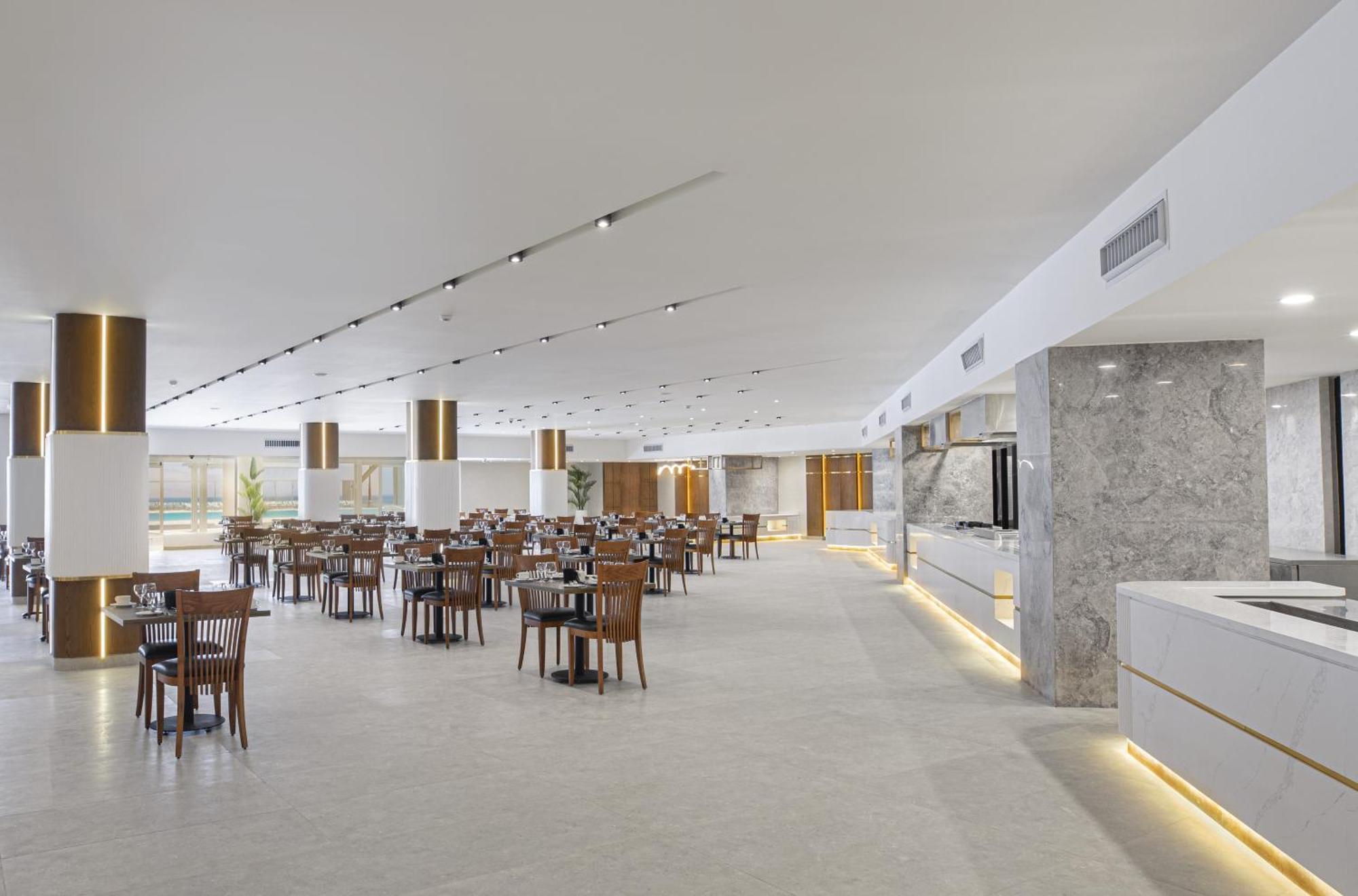 V Hotel Sharm El Shiekh Exterior photo The photo shows a modern restaurant interior. It features a spacious, well-lit environment with a clean design. The flooring is light-colored, and the walls have a minimalist aesthetic. There are several wooden tables and chairs arranged neatly throu