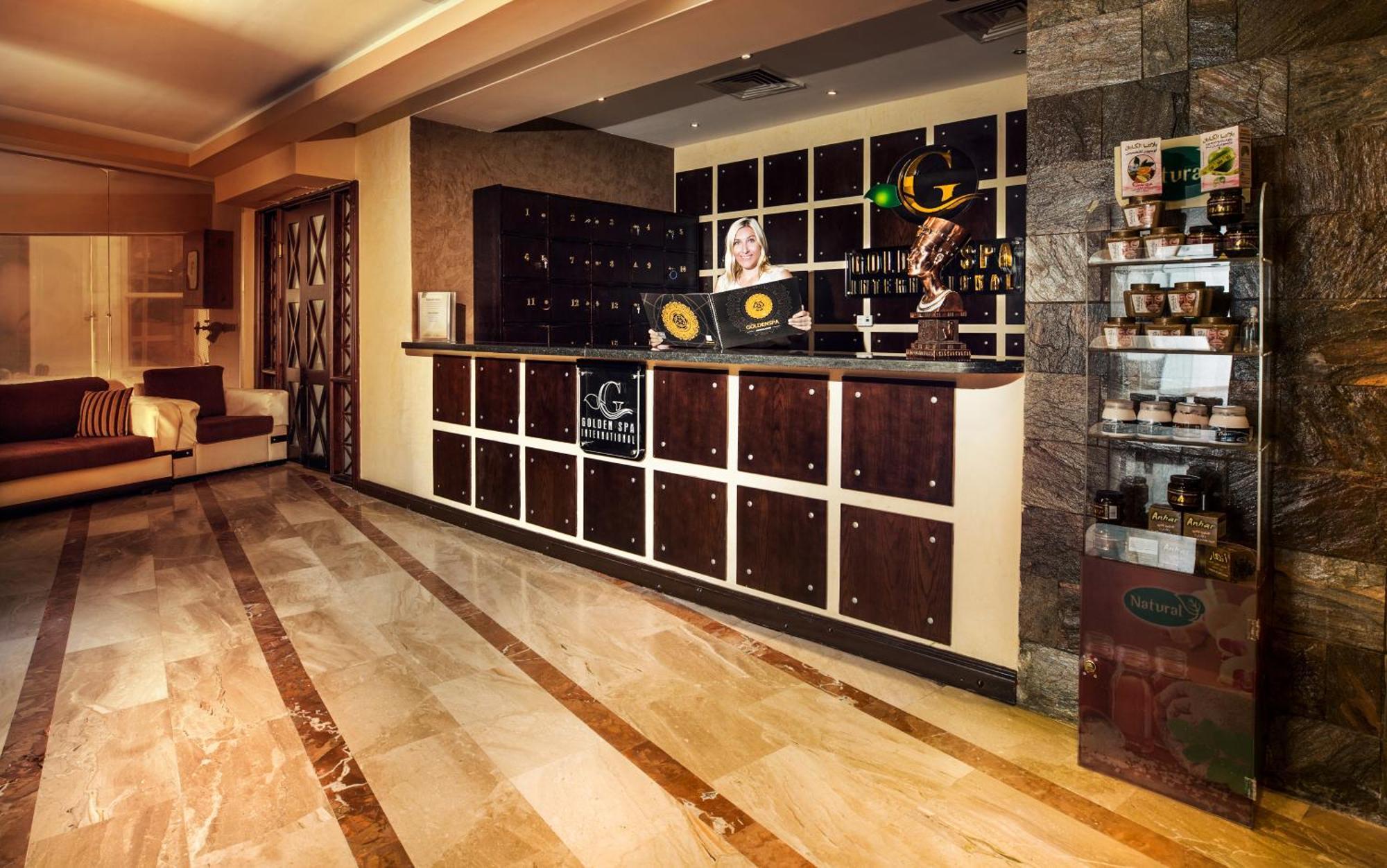 V Hotel Sharm El Shiekh Exterior photo The photo shows the interior of a hotel lobby or reception area. In the foreground, there is a reception desk made of dark wood with a grid-like pattern of storage compartments. Behind the desk, there appears to be a receptionist with a friendly deme