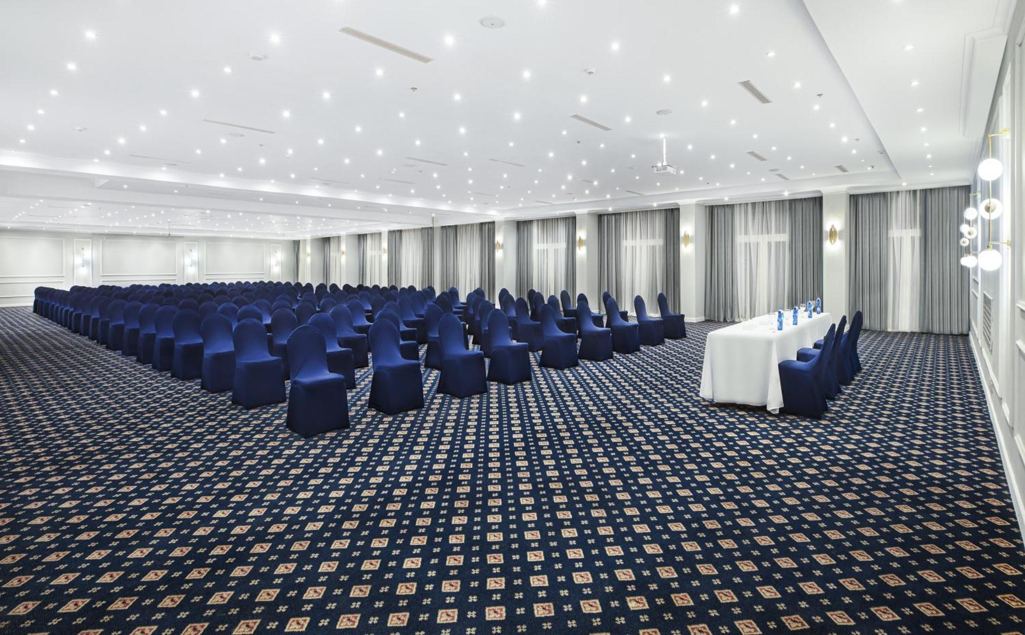 V Hotel Sharm El Shiekh Exterior photo The photo shows a spacious conference room or event hall. It features rows of blue chairs arranged neatly in a grid pattern, all facing towards a front area where there is a long, white table. The room is well-lit with bright overhead lights and has 
