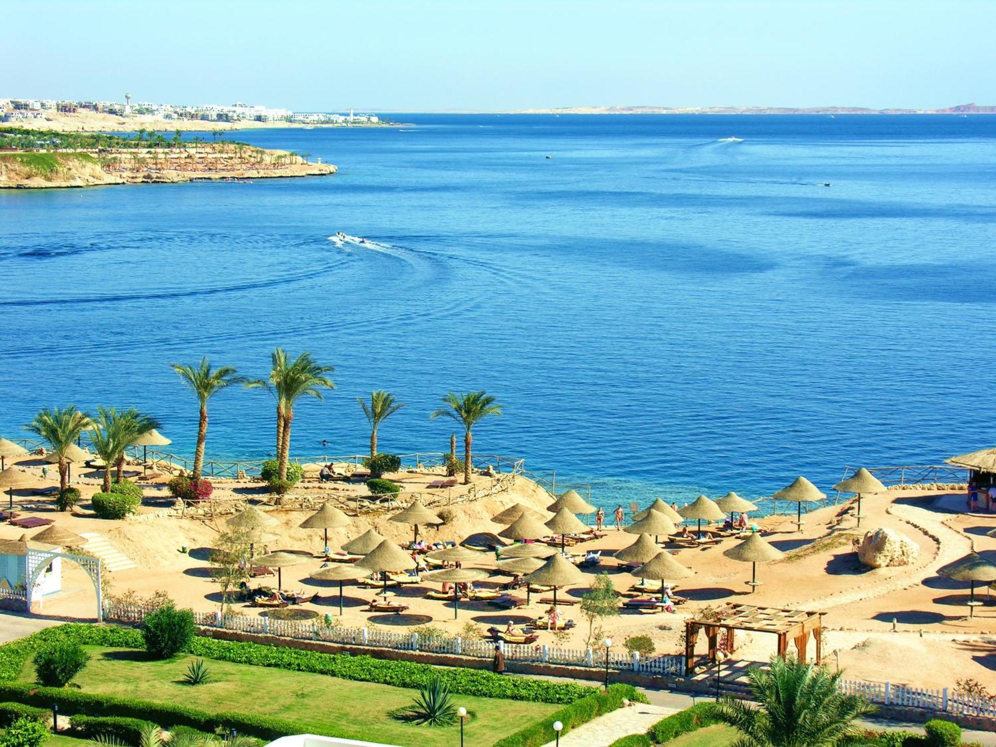 V Hotel Sharm El Shiekh Exterior photo The photo depicts a beautiful coastal scene featuring a serene blue sea and a sandy beach. There are several thatched umbrellas set up along the beach, suggesting a relaxing atmosphere for sunbathing or lounging. Palms are scattered throughout the ar