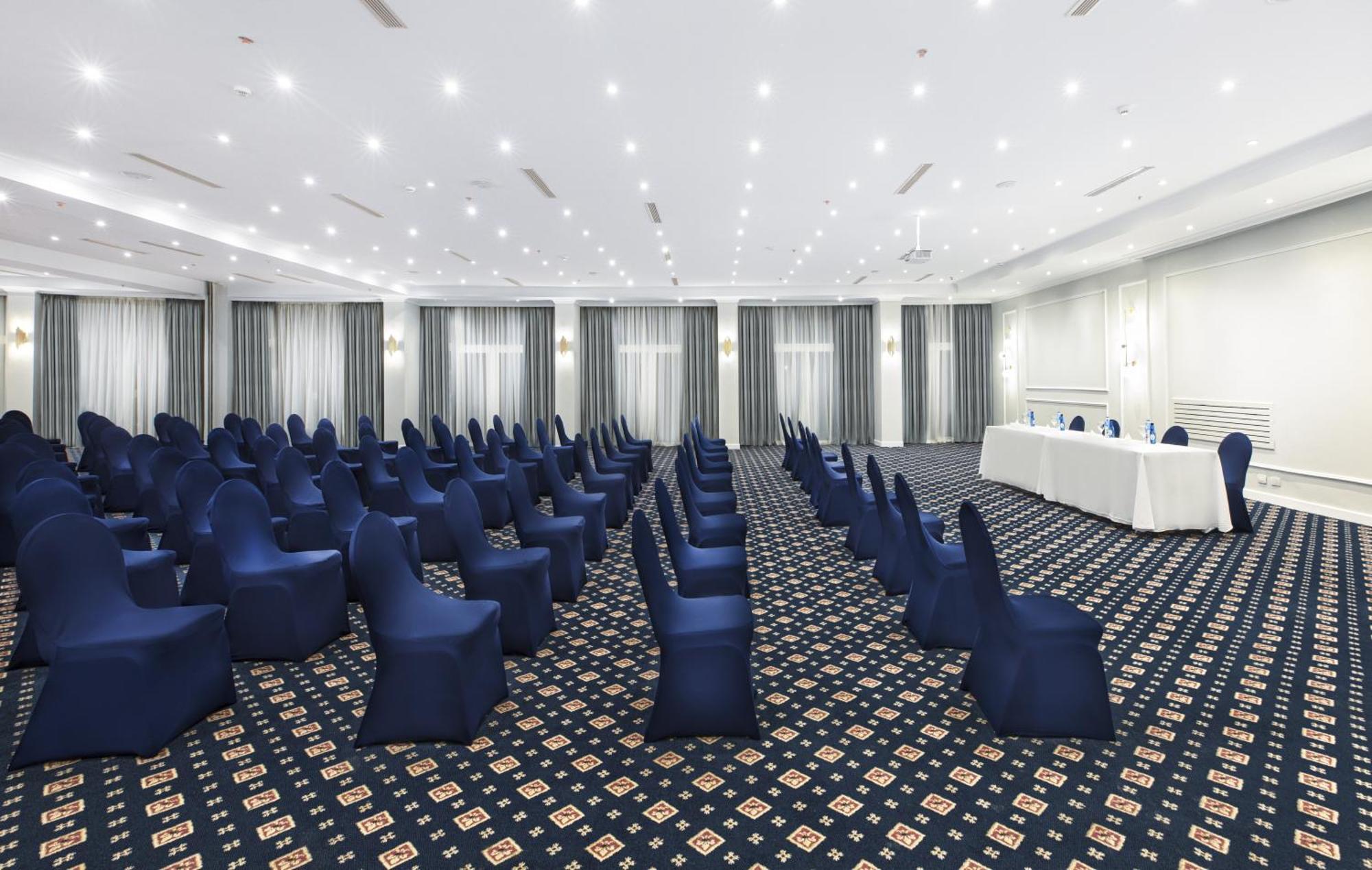 V Hotel Sharm El Shiekh Exterior photo The photo shows a spacious conference room set up for an event or meeting. There are several rows of blue chairs, each covered with blue chair covers, facing towards a long table at the front of the room. The table is covered with a white tablecloth 